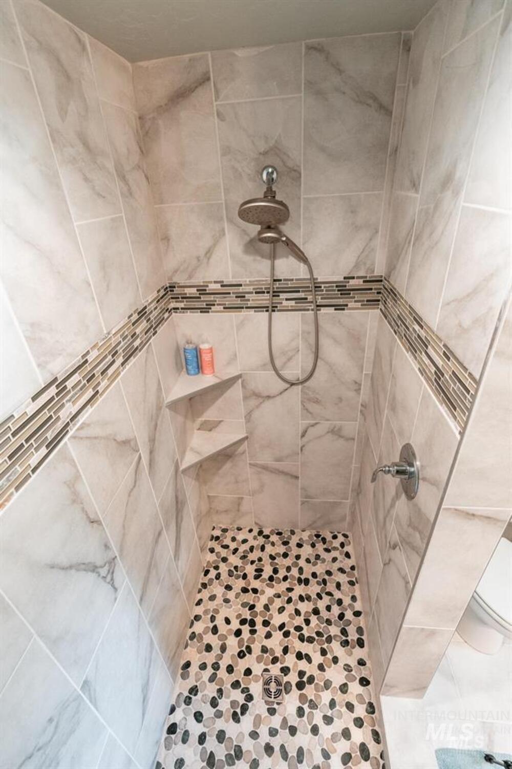 21 Melonie Loop Cascade, ID 83611 - Photo 18 of 27 Walk-in shower featuring large format wall tiles, a decorative mosaic tile band, a rainfall showerhead with a handheld sprayer, built-in shelving, and a pebble-tiled floor