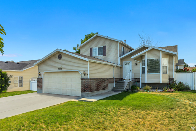5529 Ridge Hollow Way Kearns, UT 84118 - Photo 2 of 23 Split-level residence featuring light-colored siding, a two-car garage with a panel door, and brick detailing on the lower exterior