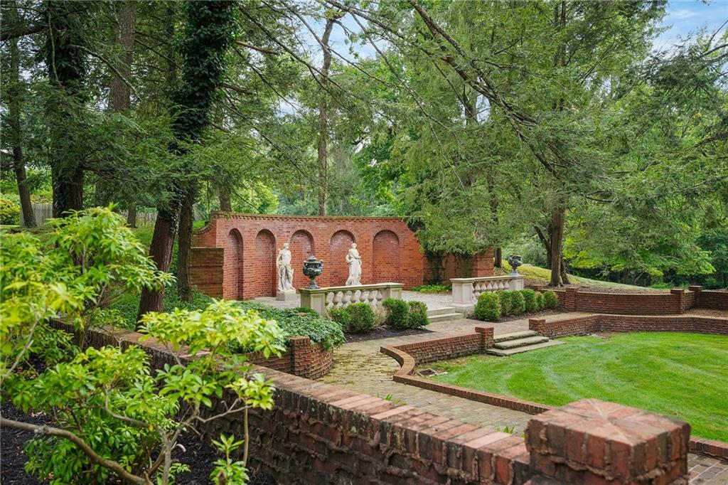 898 Persimmon Road Sewickley, PA 15143 - Photo 5 of 50 This elegant garden scene features a curved red brick wall with a series of arched niches showcasing classical statues. Stone balustrades and urns add architectural charm, while neatly manicured hedges and tiered brick pathways lead to a lush green lawn, all framed by mature trees for a serene, secluded atmosphere.