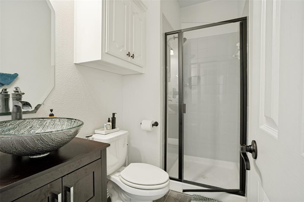 1461 Ramsbury Lane Prosper, TX 75078 - Photo 17 of 30 Bathroom featuring a vessel sink with a dark wood-finish vanity, an octagonal mirror, a toilet, a glass-enclosed shower with black framing, and white cabinetry