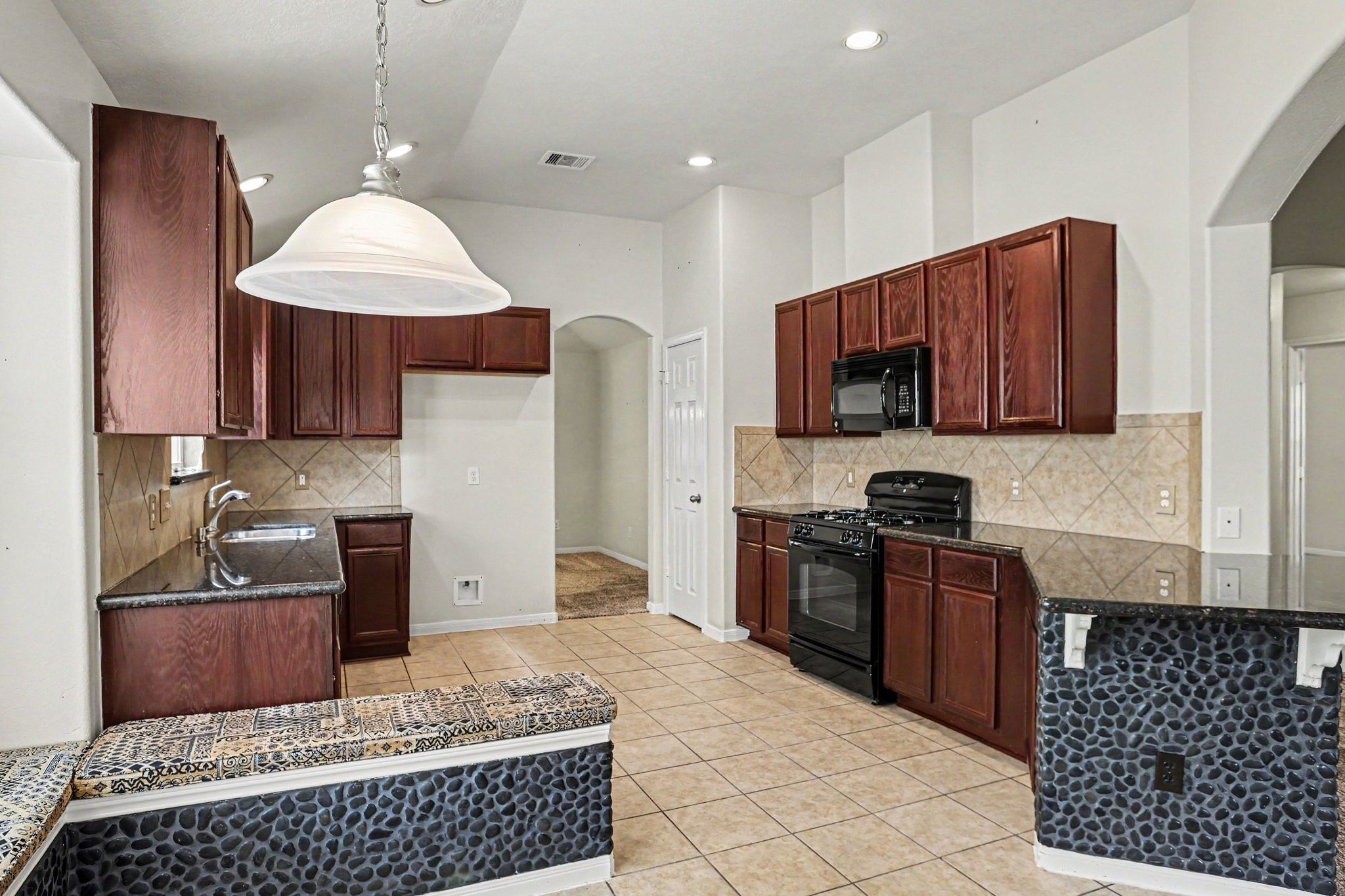 25222 Ginger Ranch Drive Katy, TX 77494 - Photo 7 of 33 This kitchen features elegant dark wood cabinetry, granite countertops, and a tile backsplash. It includes black appliances with a gas range and oven. The space is well-lit with recessed lighting and a large pendant lamp, complementing the tile flooring. An archway leads to other areas of the home.