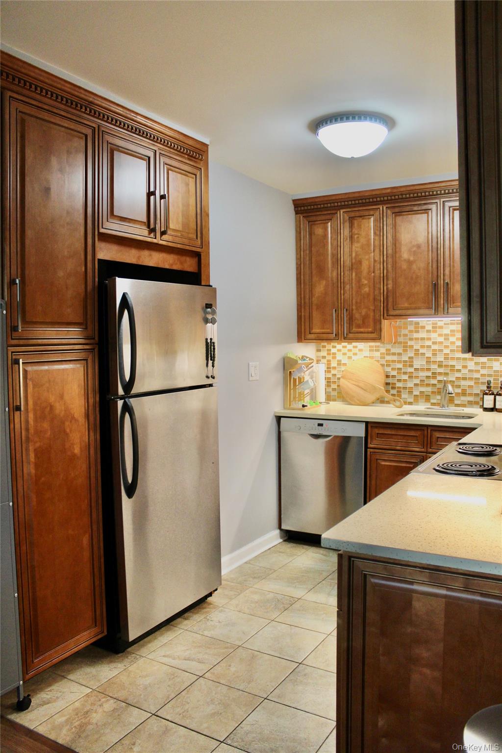 7-24 166th Street, Unit 5D Queens, NY 11357 - Photo 3 of 21 Kitchen with stainless steel appliances, backsplash, light tile patterned flooring, light stone counters, and brown cabinets