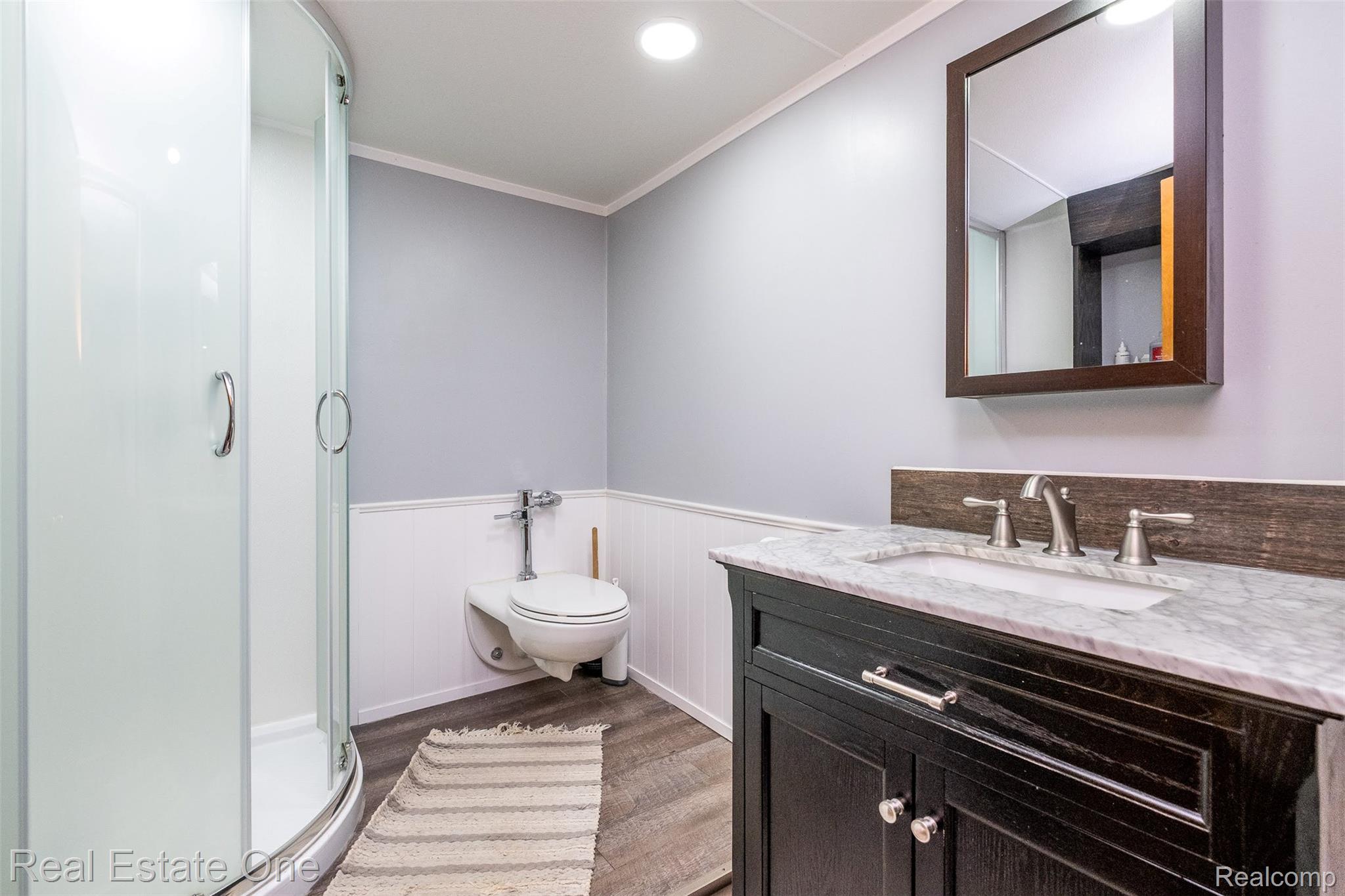 11030 Marshall Road South Lyon, MI 48178 - Photo 58 of 83 Corner shower enclosure, wall-mounted toilet, wood-finish flooring, dark wood vanity with white countertop, and rectangular wall mirror