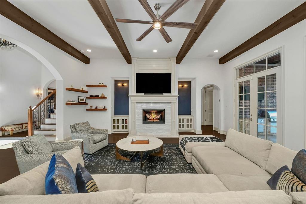 12114 Prestonridge Road Dallas, TX 75230 - Photo 17 of 40 Living room featuring exposed ceiling beams, a white stacked stone fireplace with a mantel, built-in shelving, French doors, and an arched entry to the staircase