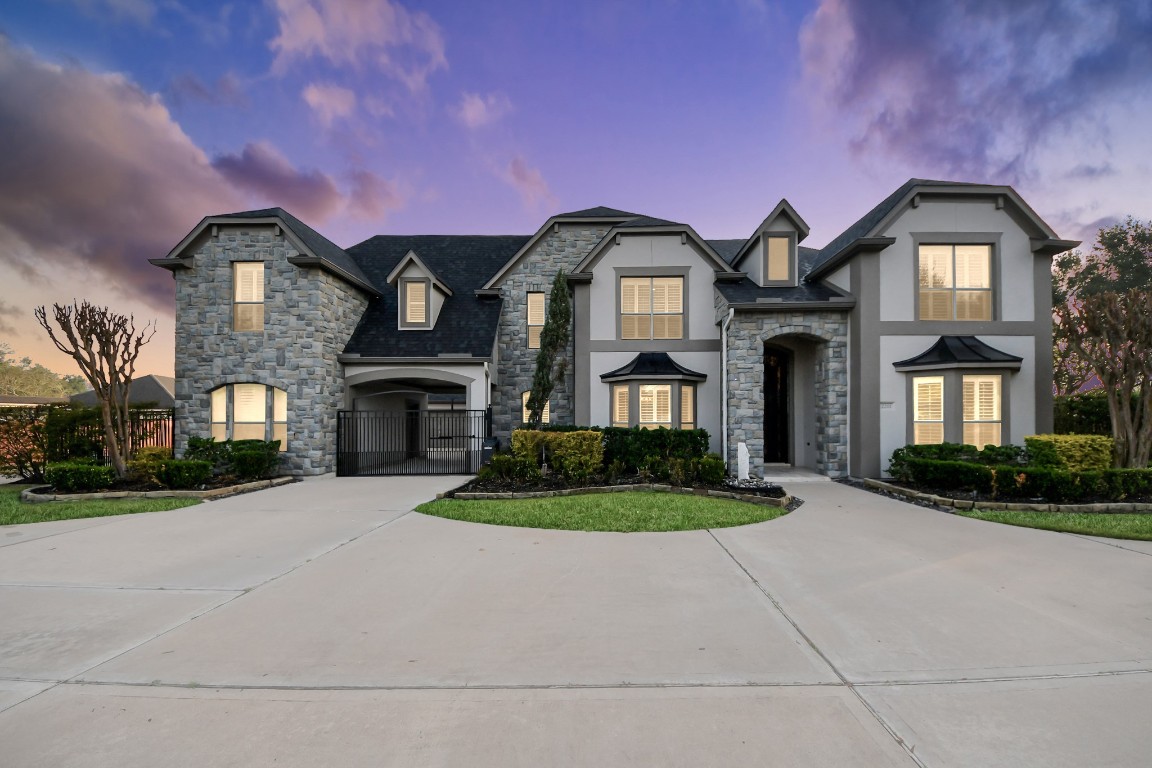 This stunning two-story home features a blend of stone and stucco exterior, with elegant architectural details and a spacious driveway leading to a gated entry. The well-maintained landscaping enhances the curb appeal, set against a picturesque evening sky.