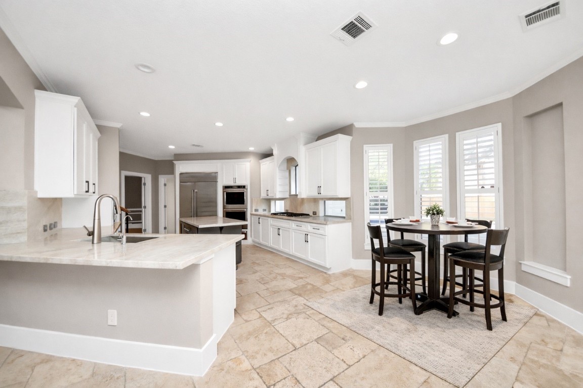 2211 Braer Ridge Drive Katy, TX 77494 - Photo 11 of 50 This modern kitchen features sleek white cabinetry, a spacious island with a sink, and stainless steel appliances. The open layout includes a cozy dining area with a round table set near large windows, allowing for ample natural light. The neutral tones and tiled flooring create a warm, inviting atmosphere.
