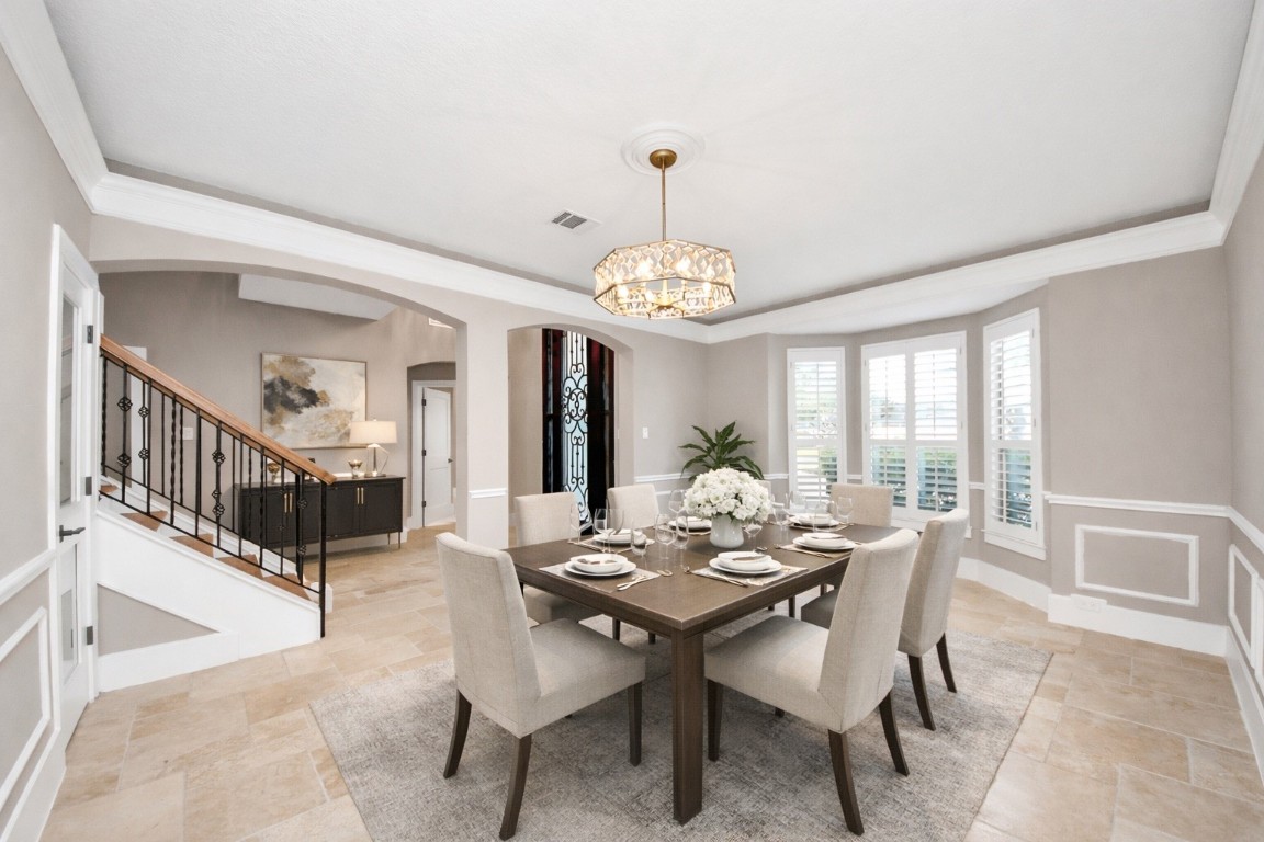 2211 Braer Ridge Drive Katy, TX 77494 - Photo 10 of 50 This photo showcases an elegant dining room with a large wooden table, stylish chandelier, and ample natural light from bay windows with plantation shutters. The space features neutral tones, wainscoting, and an adjacent staircase with wrought iron accents.