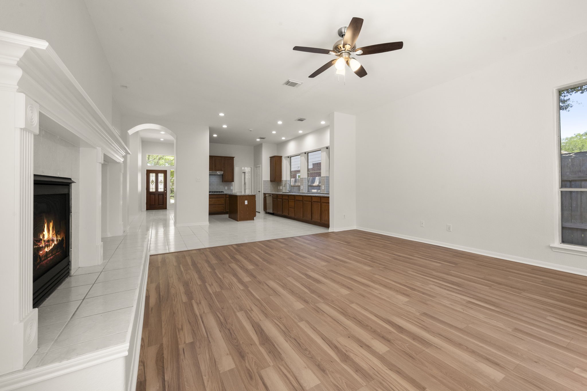 2018 Copperwood Park Lane Spring, TX 77386 - Photo 23 of 33 Spacious open-concept living area with a cozy gas fireplace, hardwood flooring, and a ceiling fan. The area flows into a modern kitchen with wooden cabinetry and ample natural light from large windows.
