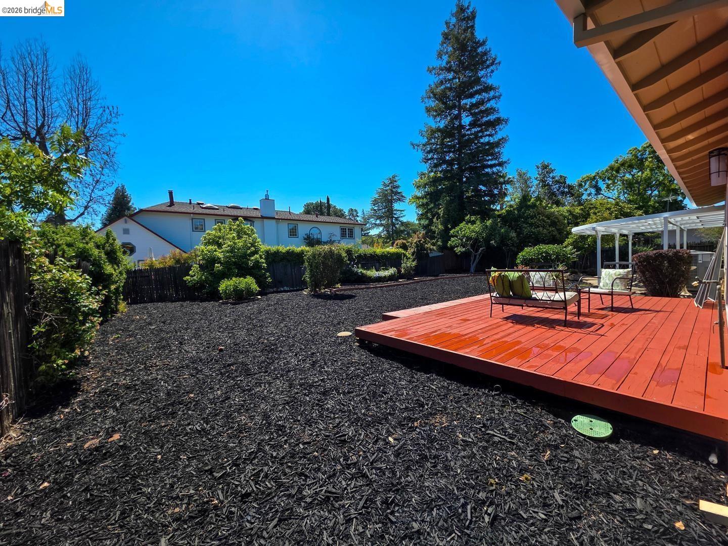 965 Springer Road Los Altos, CA 94024 - Photo 21 of 31 Expansive backyard featuring a vibrant red-stained wooden deck, a white pergola structure, and surrounding landscaping with mature trees and dark wood chip ground cover