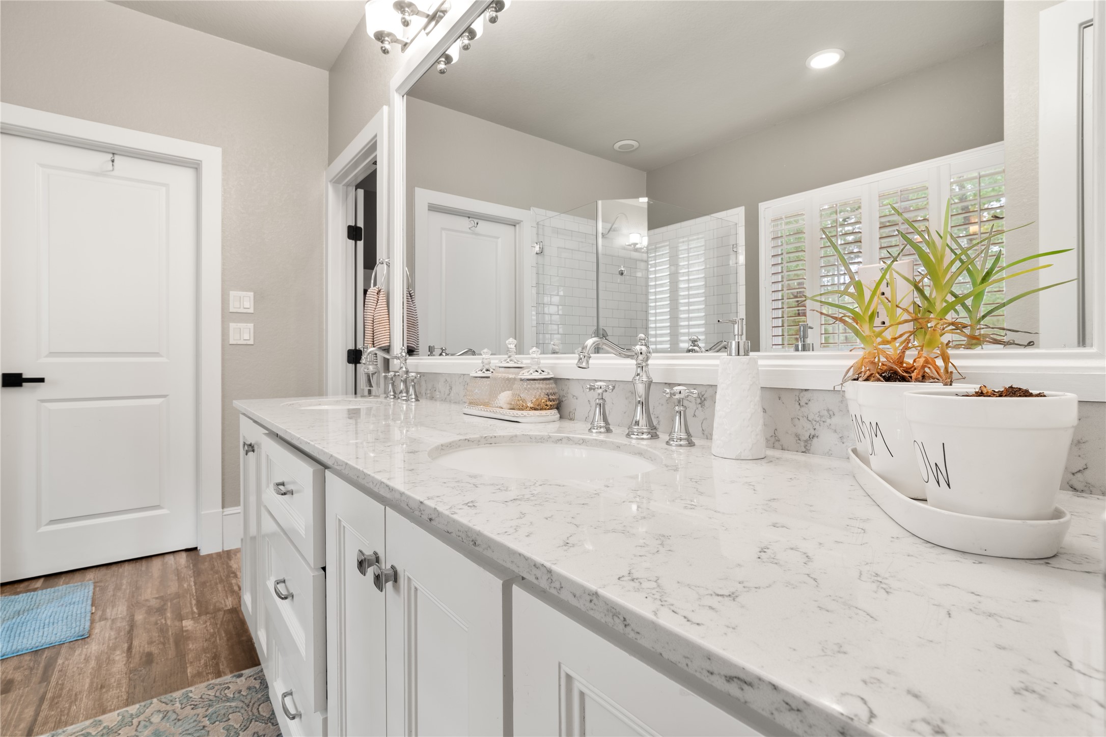 36 Wind River Drive Conroe, TX 77384 - Photo 31 of 45 This photo showcases a modern bathroom with a spacious marble countertop and dual sinks, featuring elegant fixtures. The room has a neutral color palette, wood flooring, and ample natural light from a window with white shutters. A glass-enclosed shower adds a touch of luxury.