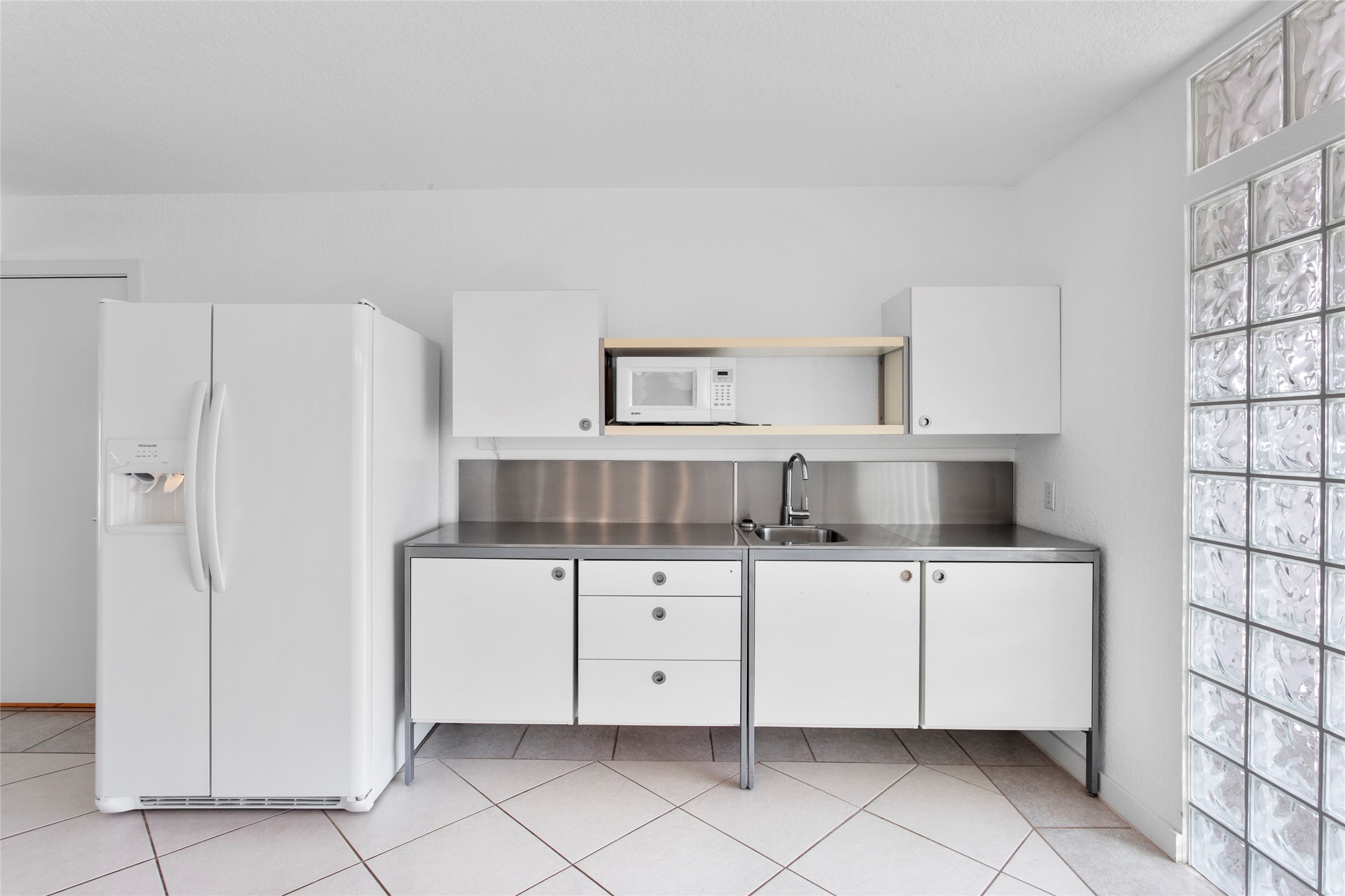 67 Harbor Lane Kemah, TX 77565 - Photo 30 of 47 This kitchen features a minimalist design with sleek white cabinets, stainless steel countertops, microwave and ample storage, complemented by a glass block wall for natural light.