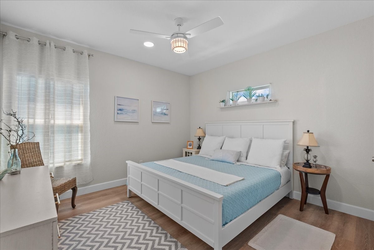 130 Beachcomber Avenue Surfside Beach, TX 77541 - Photo 17 of 33 This bedroom features a cozy, bright design with a large window, light neutral walls, and wooden flooring. It includes a white bed with blue bedding, a ceiling fan, decorative wall art, and a small shelf with plants. A side table with lamps and a dresser add functionality and charm.