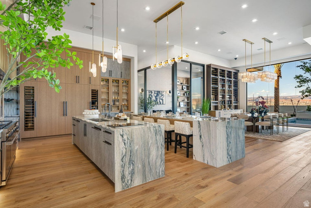 2867 East Granite Way St. George, UT 84790 - Photo 11 of 69 Open-concept kitchen and dining area featuring wood-finish flooring, two large stone islands, custom light wood cabinetry, and a full-height wine storage display