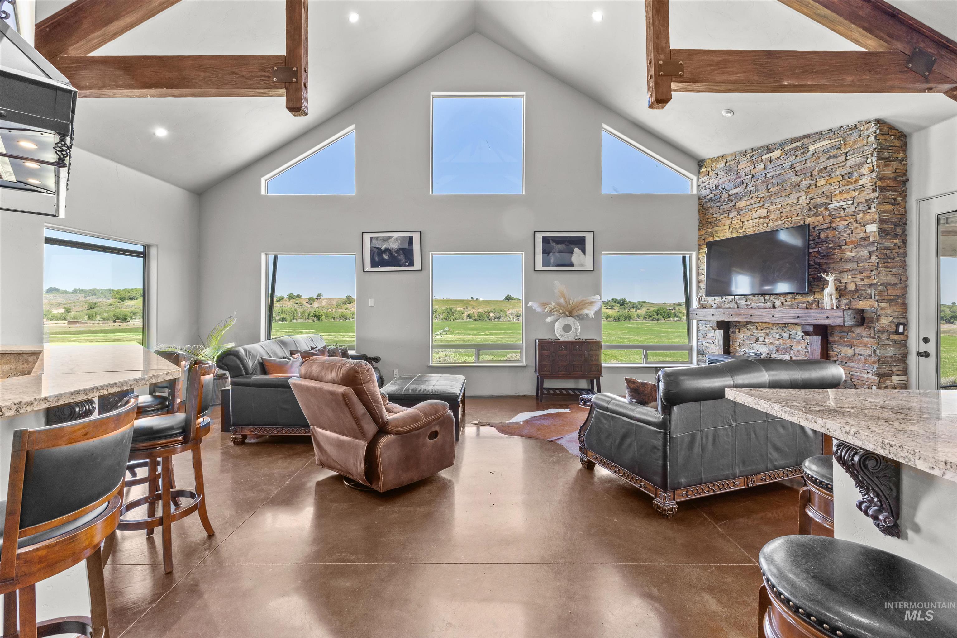 1030 Desert Glen Road Adrian, OR 97901 - Photo 32 of 47 Spacious living area featuring a vaulted ceiling with exposed wood beams, a stacked stone fireplace, large windows providing expansive outdoor scenery, polished concrete flooring, and a stone-look kitchen island with bar seating