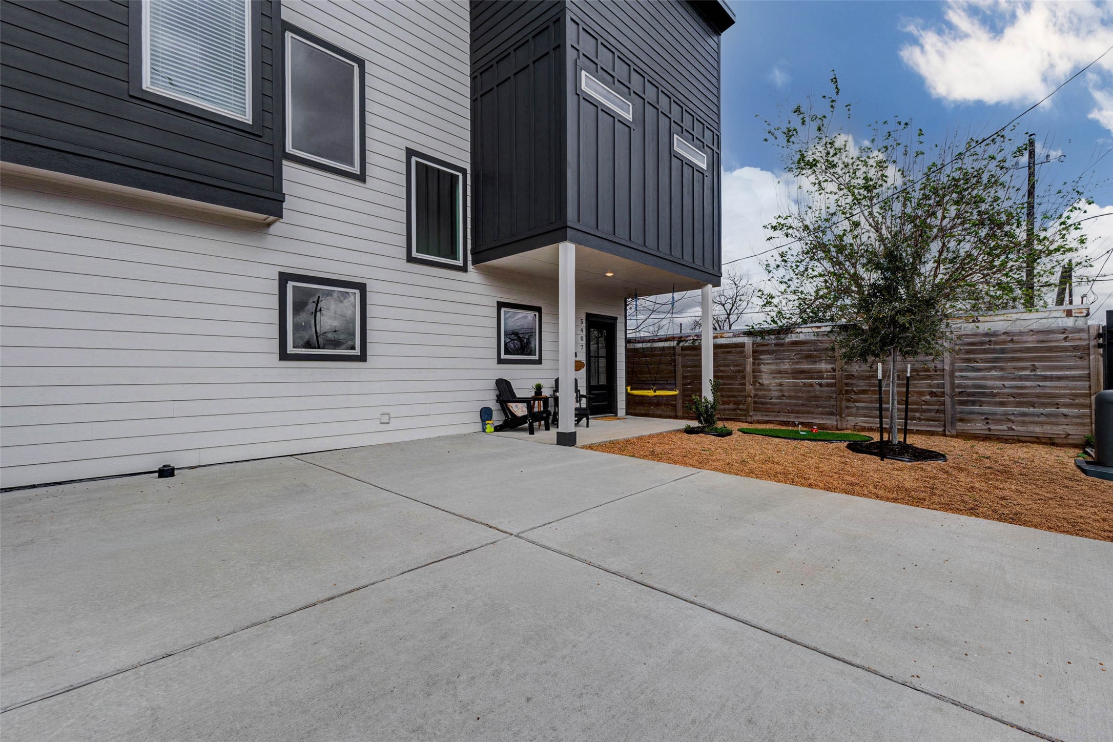 5407 Lee Street Houston, TX 77020 - Photo 17 of 17 This photo showcases a modern home's exterior with a clean, minimalist design. It features a spacious concrete patio, ideal for outdoor activities, and a small garden area with young trees. The home's siding combines white and dark panels for a contemporary look, and a wooden fence offers privacy.