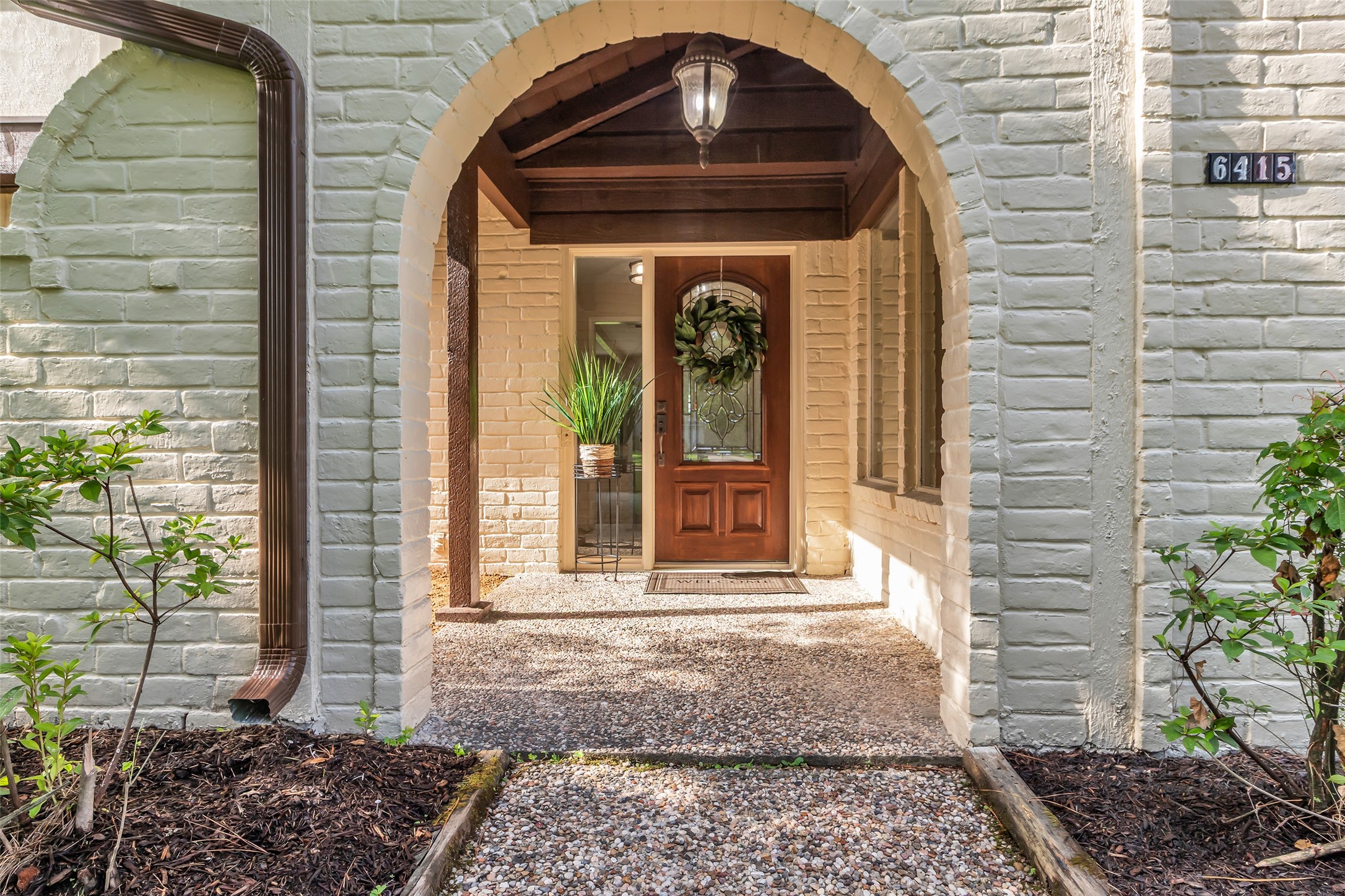 Arched brick accents This inviting entrance features a charming brick archway leading to a wooden front door with decorative glass. The pathway is lined with greenery, and a welcoming light fixture enhances the cozy ambiance.