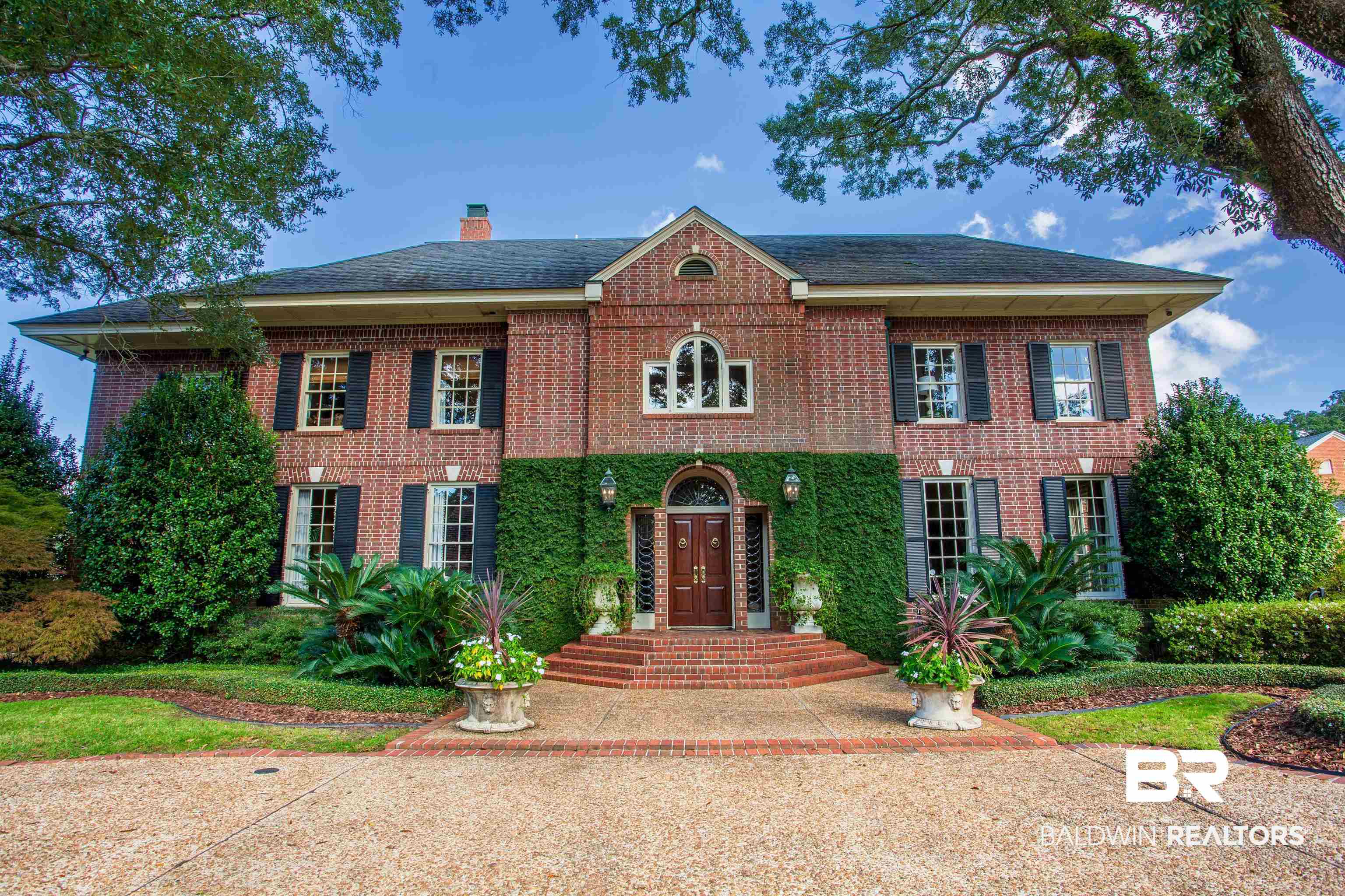 150 Austill Avenue Mobile, AL 36608 - Photo 4 of 39 Traditional brick exterior featuring a prominent gabled entry, symmetrical window placement with dark shutters, established landscaping with manicured shrubs, a brick-paved walkway, and a large paved driveway