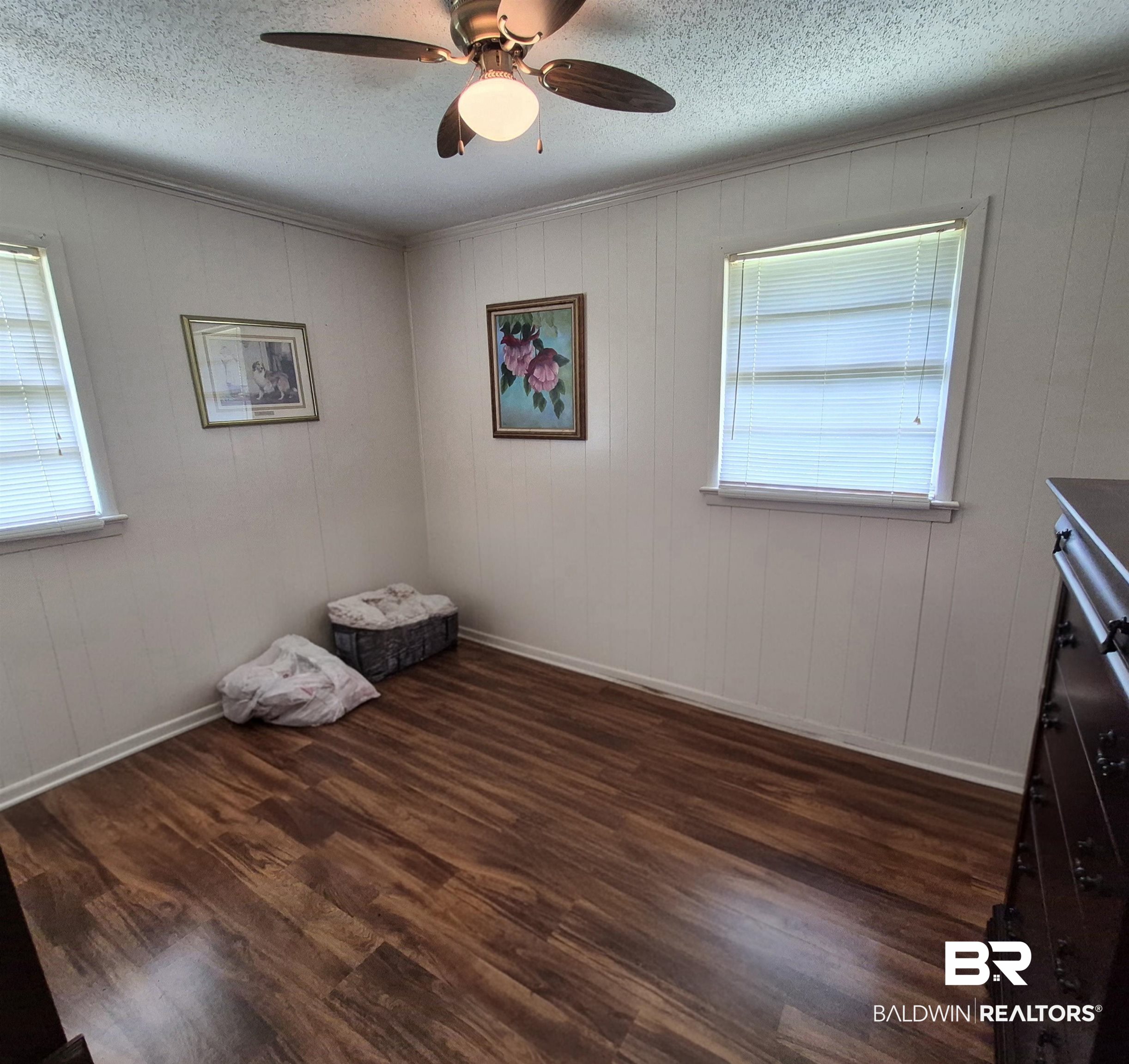 108 Azalea Street Evergreen, AL 36401 - Photo 12 of 19 Room featuring wood-finish flooring, two windows with blinds, a ceiling fan with integrated lighting, and vertical paneling on the walls