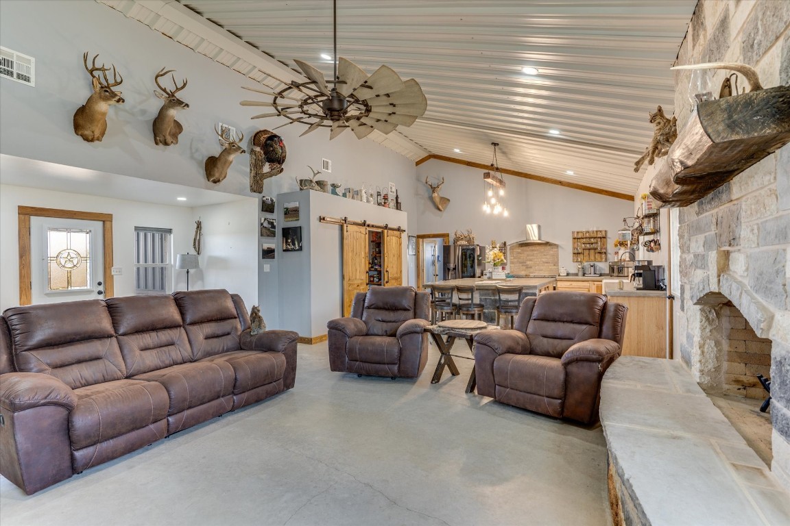 Living room with a barn door, a stone fireplace, ceiling fan, wooden ceiling, and high vaulted ceiling