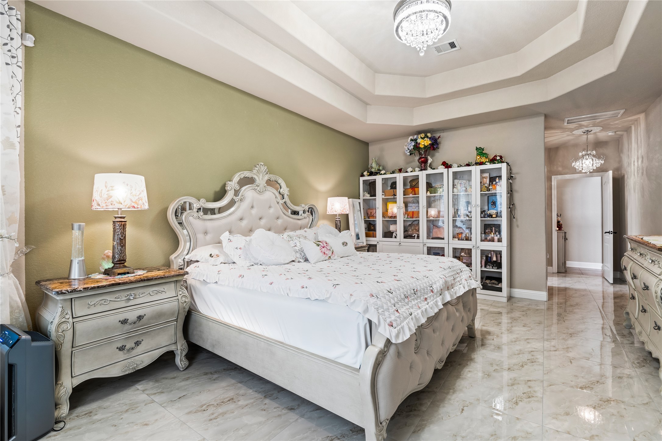 5802 Layton Meadows Lane Spring, TX 77379 - Photo 21 of 42 Master bedroom - Stunning tray ceiling with crystal chandelier, sage green accent wall, ornate French Provincial bed frame, marble tile floors, glass display cabinets, and a hallway leading to the closet/bath.