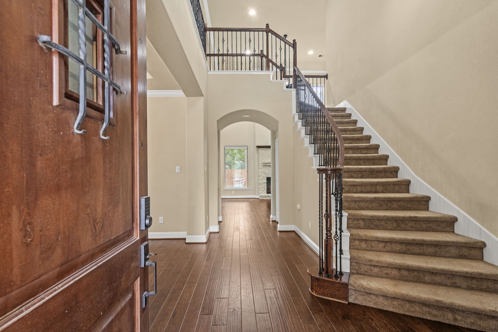 14215 Spindle Arbor Road Cypress, TX 77429 - Photo 3 of 47 This inviting entryway features a wooden door opening to a spacious foyer with elegant hardwood flooring. A carpeted staircase with decorative iron railings ascends to the upper level, while an arched doorway leads to a bright living area. High ceilings and neutral walls enhance the open feel.