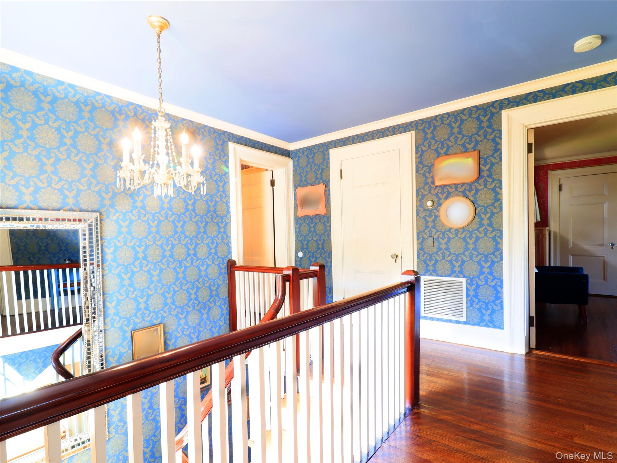11-20 Shore Road Queens, NY 11363 - Photo 33 of 45 Hardwood flooring and a classic chandelier illuminate this upper landing, featuring a white balustrade with a dark wood-finish handrail, patterned blue wallpaper, and white trim