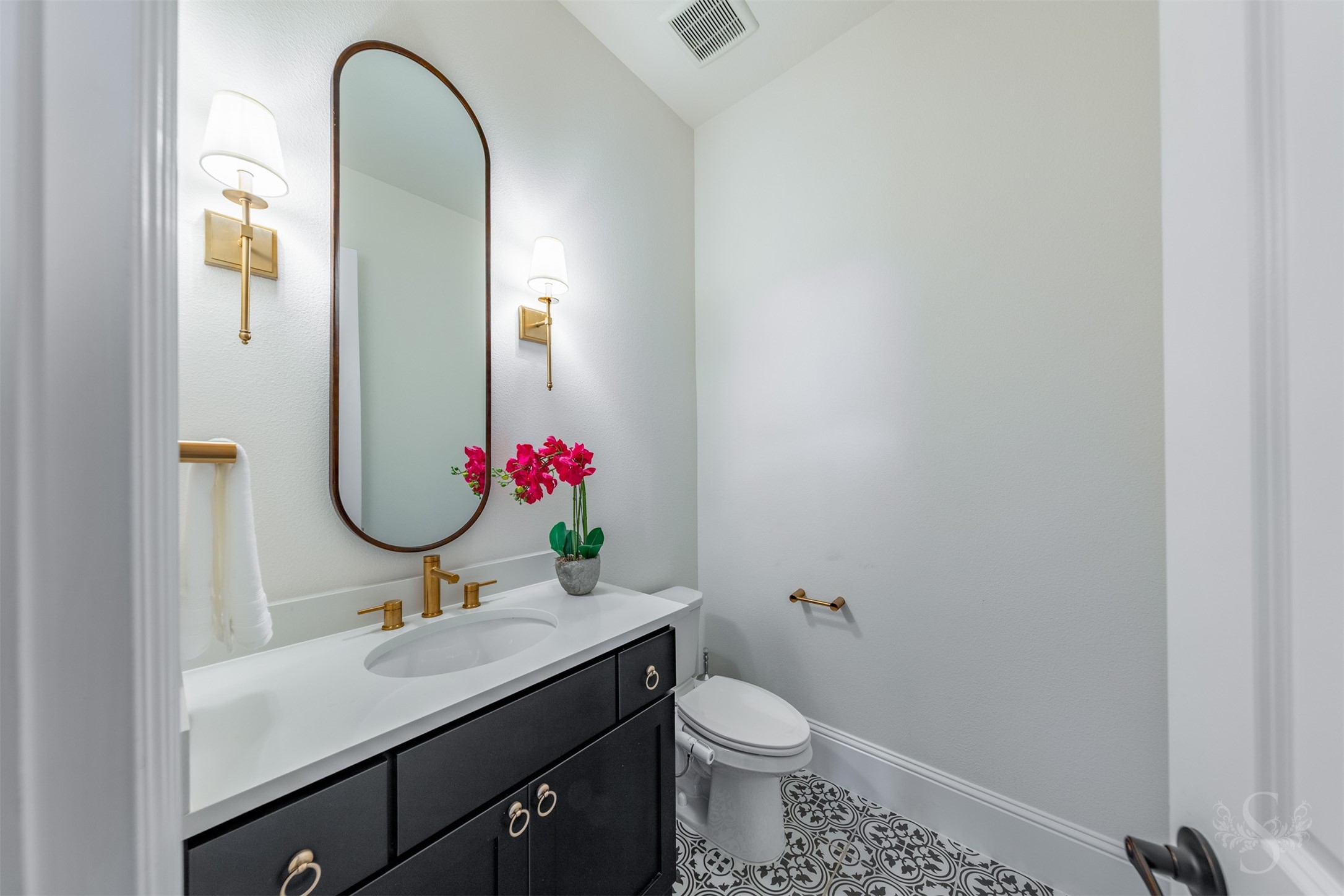 27919 Indigo Ridge Drive Fulshear, TX 77441 - Photo 33 of 50 This stylish half bath showcases designer flair with a sleek black vanity, brass hardware, and an elegant arched mirror accented by warm sconces. Patterned tile flooring adds character and charm, creating a memorable powder room for guests.
