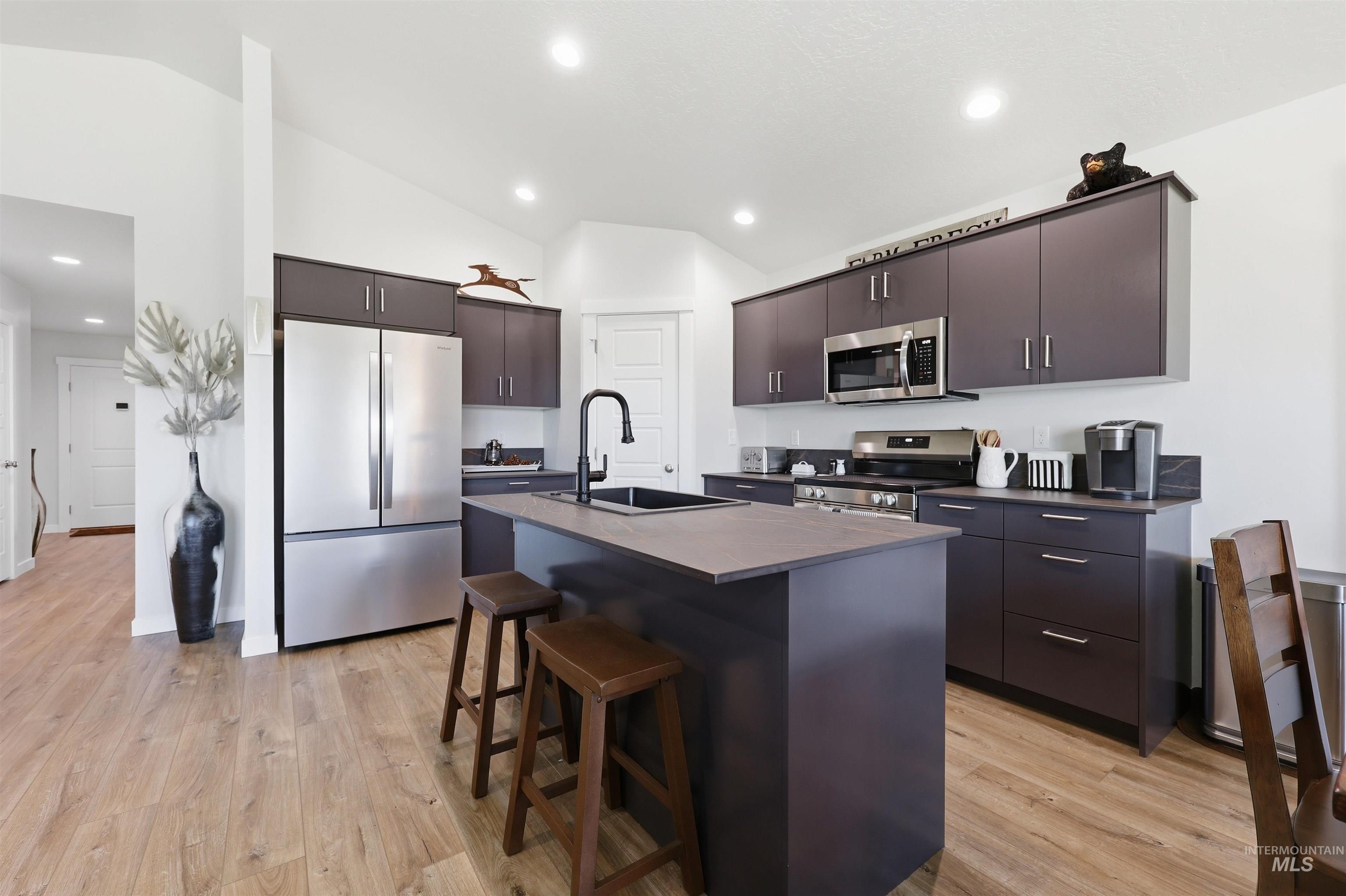3811 West Maypearl Street Meridian, ID 83642 - Photo 18 of 39 Modern kitchen featuring vaulted ceilings, wood-finish flooring, stainless steel appliances, dark flat-panel cabinetry, and an island with a black sink and faucet