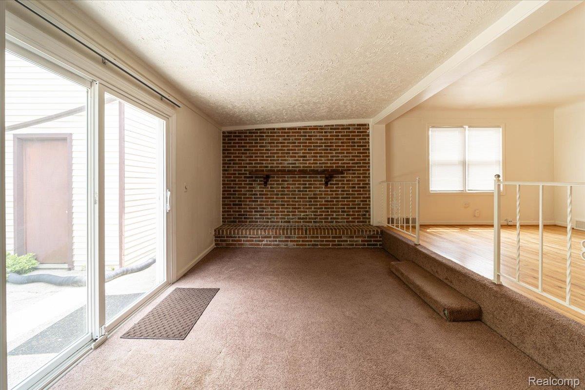 4335 Territorial Road, Unit B Rochester, MI 48306 - Photo 7 of 20 Carpeted living area featuring a brick accent wall with a wood mantel, sliding glass doors to the exterior, and a raised platform with wood-finish flooring and a white metal railing
