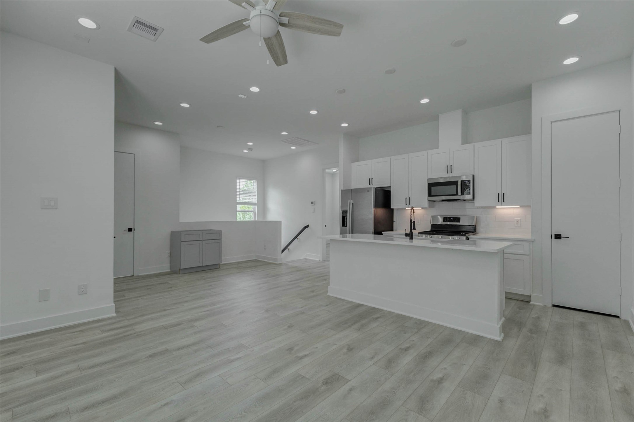 5705 Elysium Street Houston, TX 77091 - Photo 11 of 22 Expansive kitchen and living view featuring a large white island, stainless appliances, white cabinetry, black hardware, under-cabinet lighting, and light wood-look flooring. Tall ceilings, recessed lights, and an open layout give the space a polished contemporary feel with room for dining, gathering, and comfortable daily living.