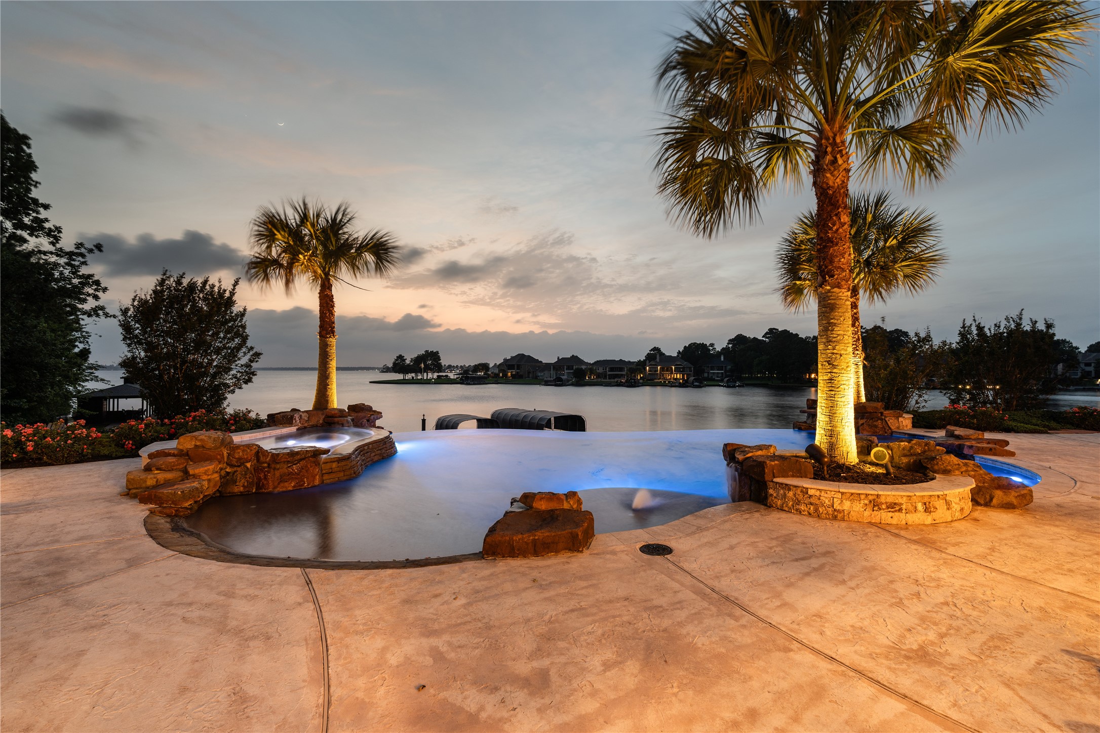 12349 Longmire Trace Conroe, TX 77304 - Photo 3 of 48 Infinity-edge pool and spa overlook the lake, framed by palm trees and glowing landscape lighting.
