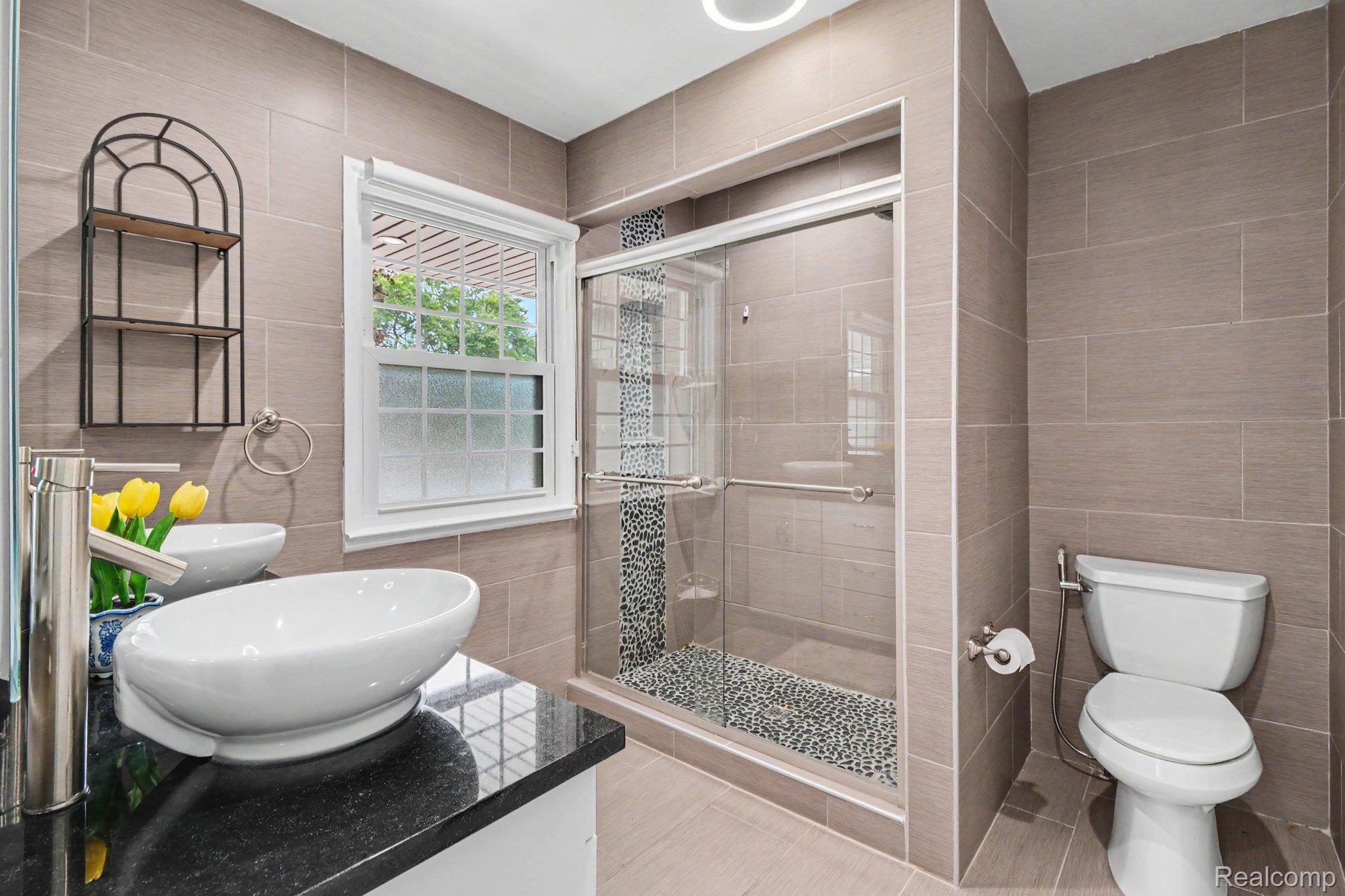 2373 Rutherford Road Bloomfield Hills, MI 48302 - Photo 17 of 28 Dual vessel sinks set on a dark countertop, walk-in shower with glass enclosure and pebble floor, and neutral wall tiling