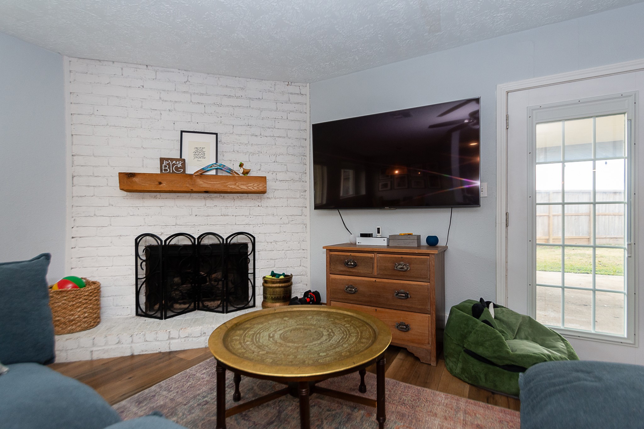 4830 Lost Oak Drive Spring, TX 77388 - Photo 16 of 41 Cozy living room with a white brick fireplace, mounted TV, and natural light from a glass door leading outside. Features a wooden mantle, stylish round table, and comfortable seating, creating a welcoming atmosphere.