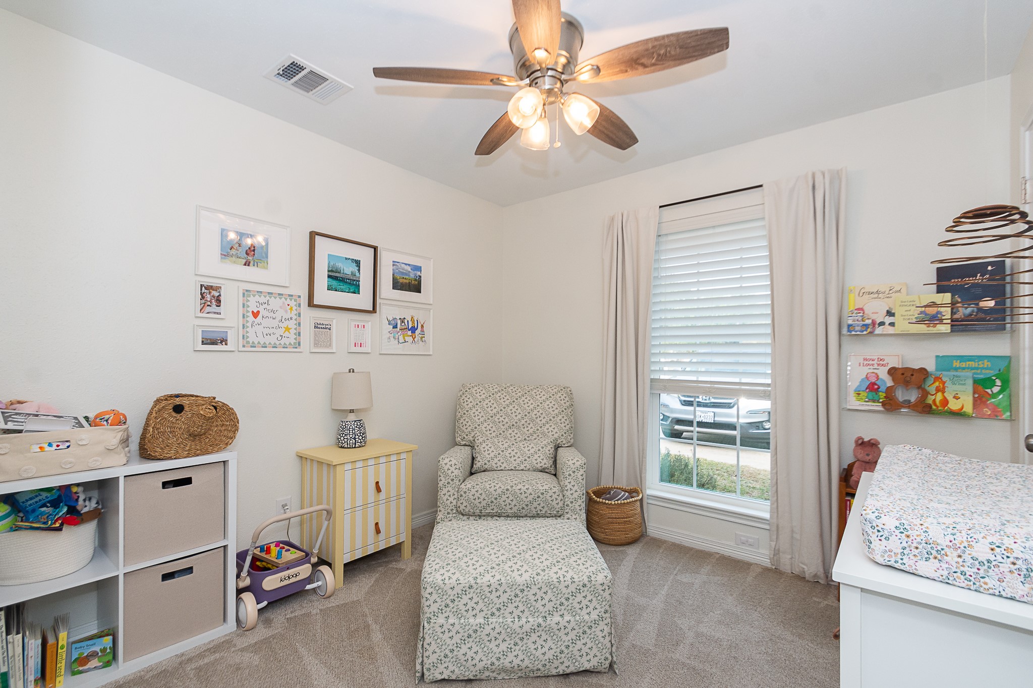 4830 Lost Oak Drive Spring, TX 77388 - Photo 19 of 41 This cozy nursery features soft carpeting, a comfortable armchair, and a ceiling fan with lights. A large window with blinds provides natural light, while wall shelves and storage units offer ample space for books and toys. The room is tastefully decorated with framed artwork and a neutral color palette.