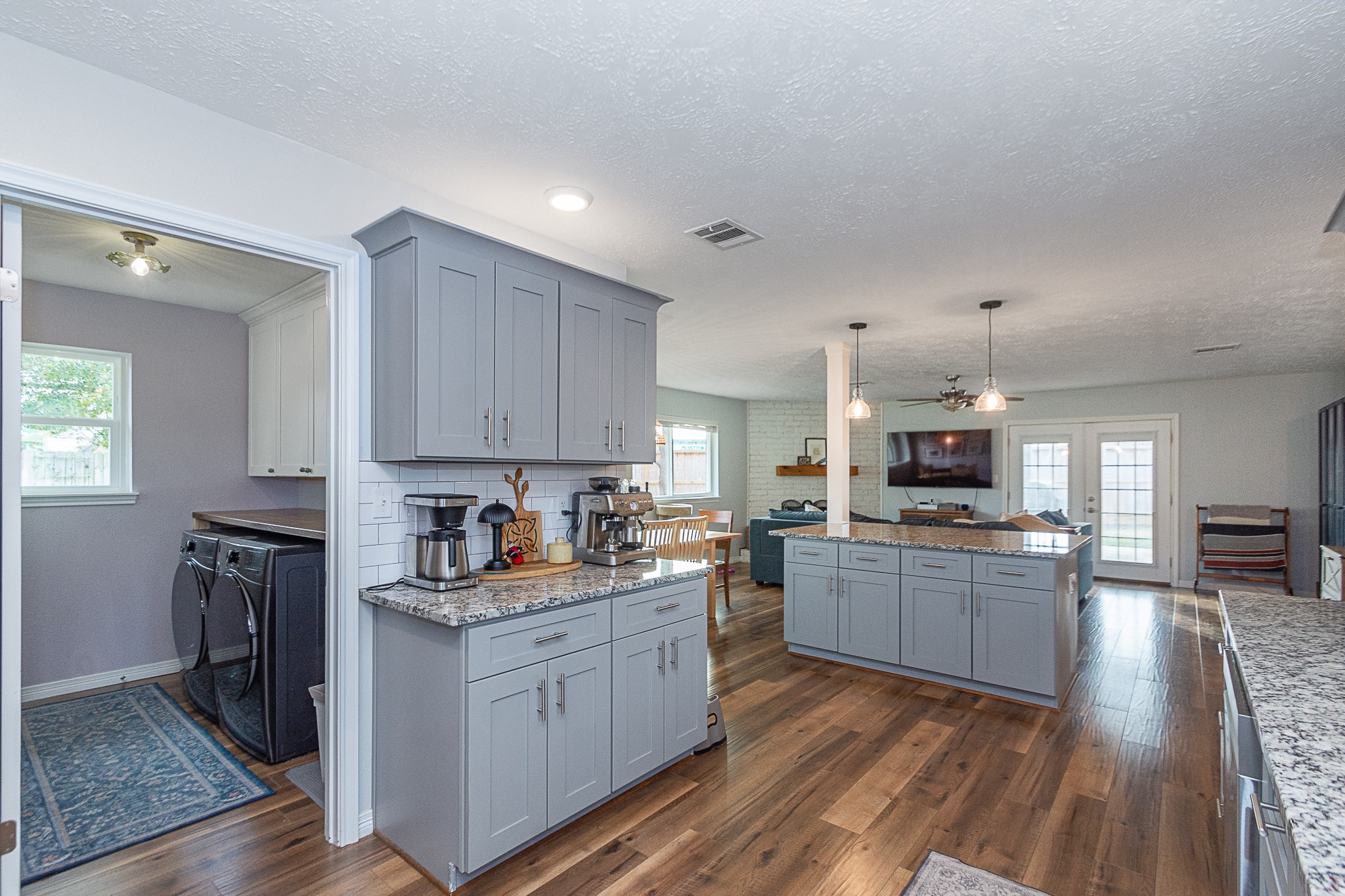 4830 Lost Oak Drive Spring, TX 77388 - Photo 6 of 41 This photo showcases an open-concept kitchen and living area with modern gray cabinetry, granite countertops, and hardwood floors. It features a spacious kitchen island with pendant lighting, and the area is well-lit with natural light from large windows and glass doors. A laundry room is conveniently adjacent to the kitchen.