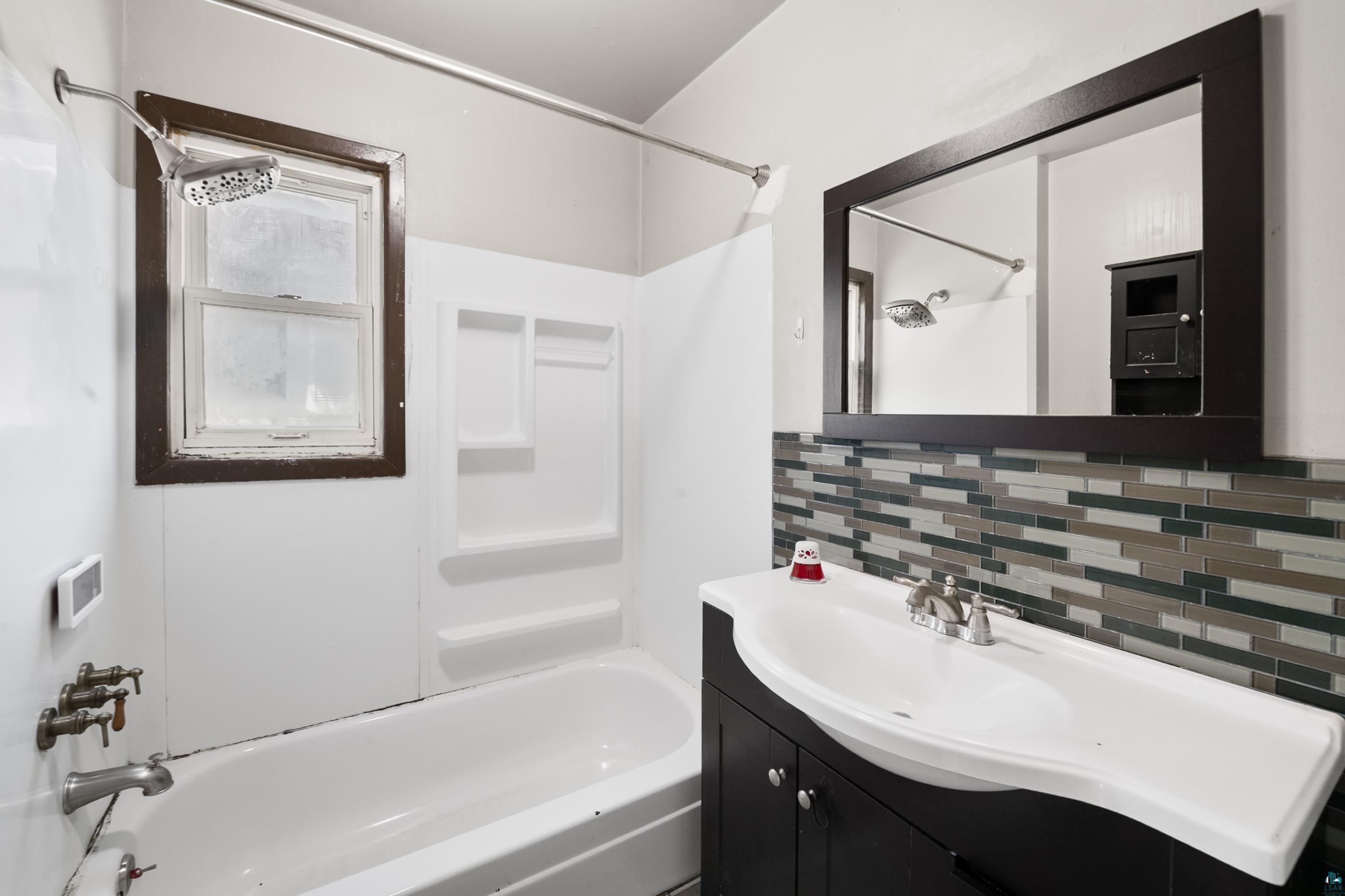 3067 Restormel Street Duluth, MN 55806 - Photo 16 of 25 Bathroom featuring a white bathtub, showerhead, window with dark trim, and a vanity with a rectangular mirror and tile backsplash