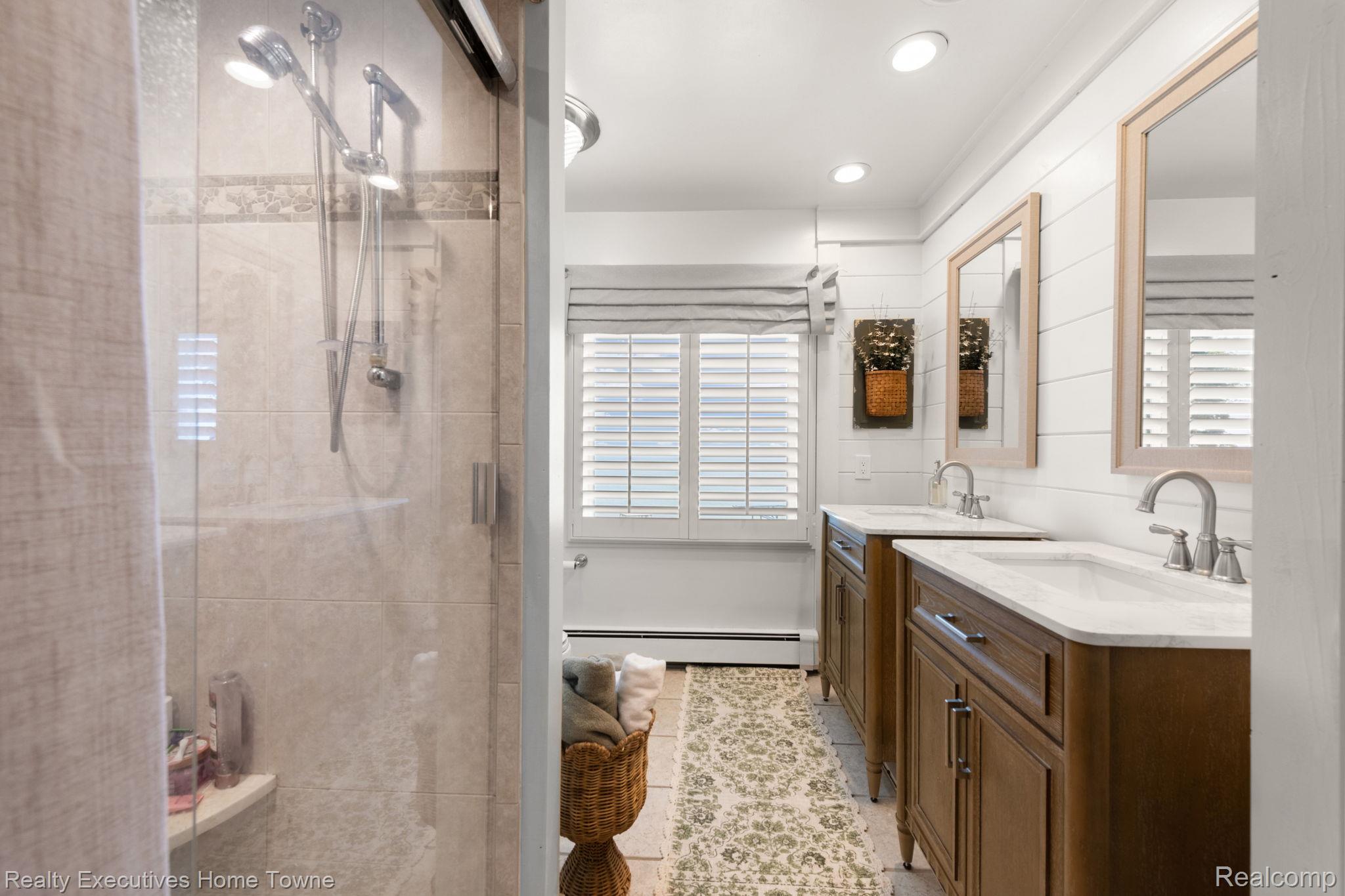 8308 Lakeshore Road Fort Gratiot, MI 48059 - Photo 61 of 79 Bathroom featuring a glass-enclosed shower with tile surround, dual wood-finish vanities with undermount sinks and light-toned countertops, two framed mirrors, and plantation shutters