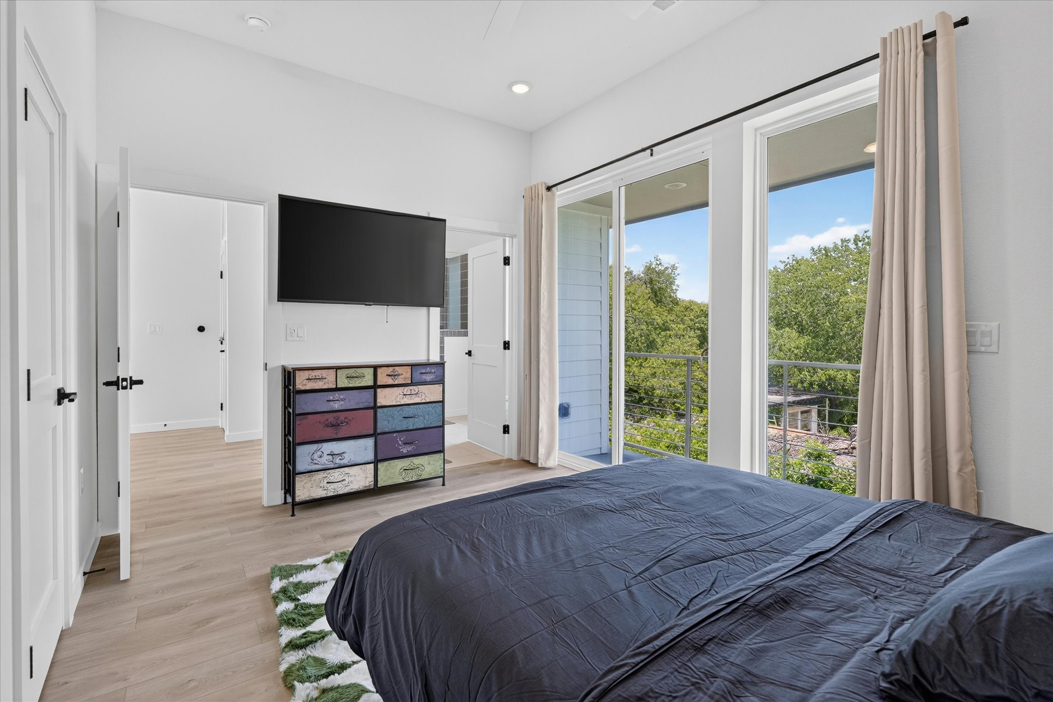 2000 Riverside Farms Road, Unit 8101 Austin, TX 78741 - Photo 16 of 41 This bedroom features a modern design with light wood flooring and a large sliding glass door that opens to a balcony with lush greenery views. It includes a wall-mounted TV and a colorful chest of drawers, offering both style and functionality.