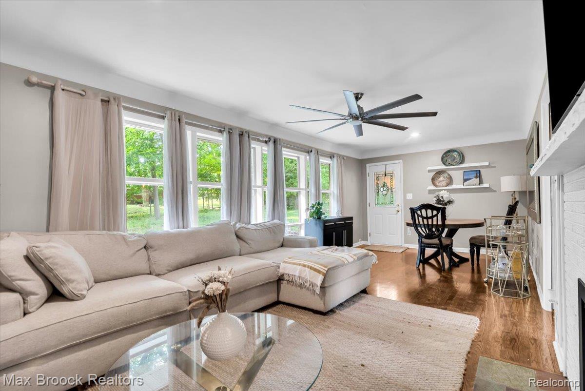 Spacious living area featuring wood-finish flooring, a contemporary ceiling fan, multiple large windows, and a classic white entry door with frosted glass