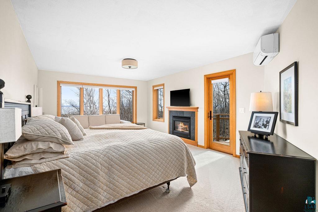 4856 Kingston Street Duluth, MN 55804 - Photo 29 of 88 Expansive bedroom featuring large windows, a built-in fireplace with a dark tile surround and wood mantel, a glass-paneled exterior door, a wall-mounted mini-split unit, and neutral carpeting