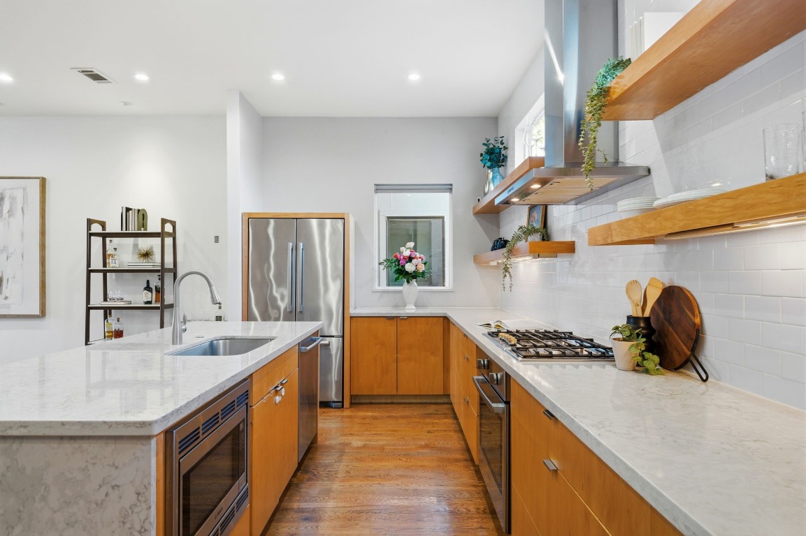 1405 Vermont Street, Unit A Houston, TX 77006 - Photo 16 of 46 Warm wood cabinetry and an oversized quartz waterfall island anchor this gourmet kitchen, featuring a professional gas cooktop and sleek built-in vent hood. Floating wood shelves and a clean subway tile backsplash provide a contemporary touch, while the open layout flows effortlessly into a spacious walk-in pantry. Hardwood floors and recessed LED lighting enhance the luxury atmosphere of this culinary centerpiece.