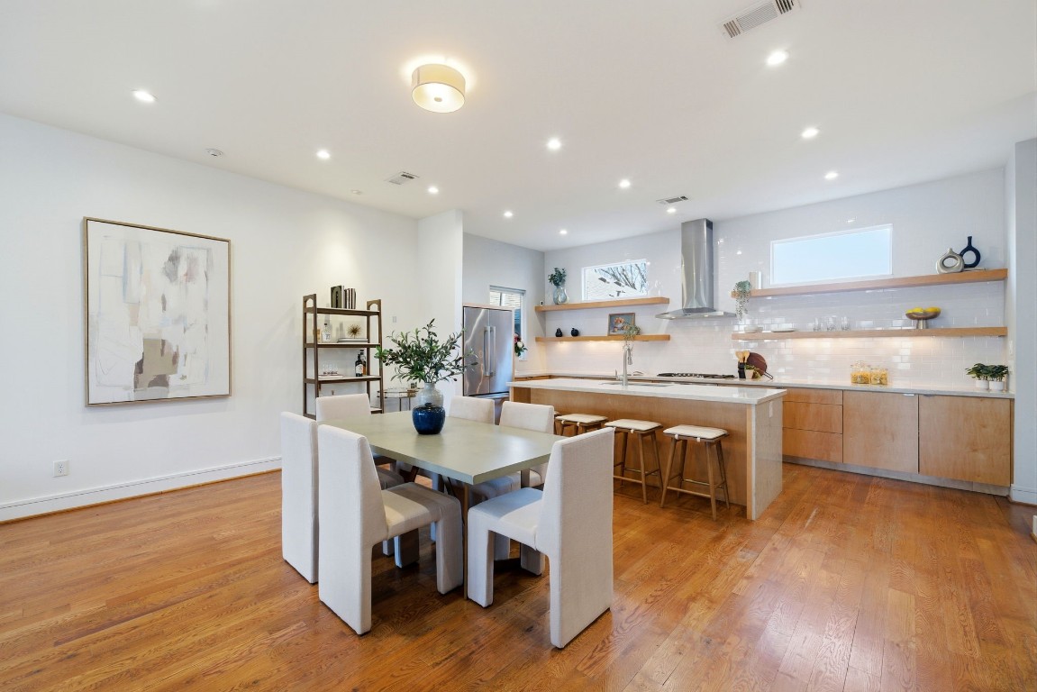 1405 Vermont Street, Unit A Houston, TX 77006 - Photo 19 of 46 Polished hardwoods and soaring ceilings unify this expansive second-floor layout, where a dedicated dining area transitions seamlessly into the gourmet kitchen. The culinary space showcases an oversized quartz waterfall island with bar seating, professional gas cooktop, and sleek built-in vent hood against a clean subway tile backsplash. Dual clerestory windows and recessed LED lighting illuminate the designer finishes, while floating wood shelves add a modern touch to the abundant cabinetry.