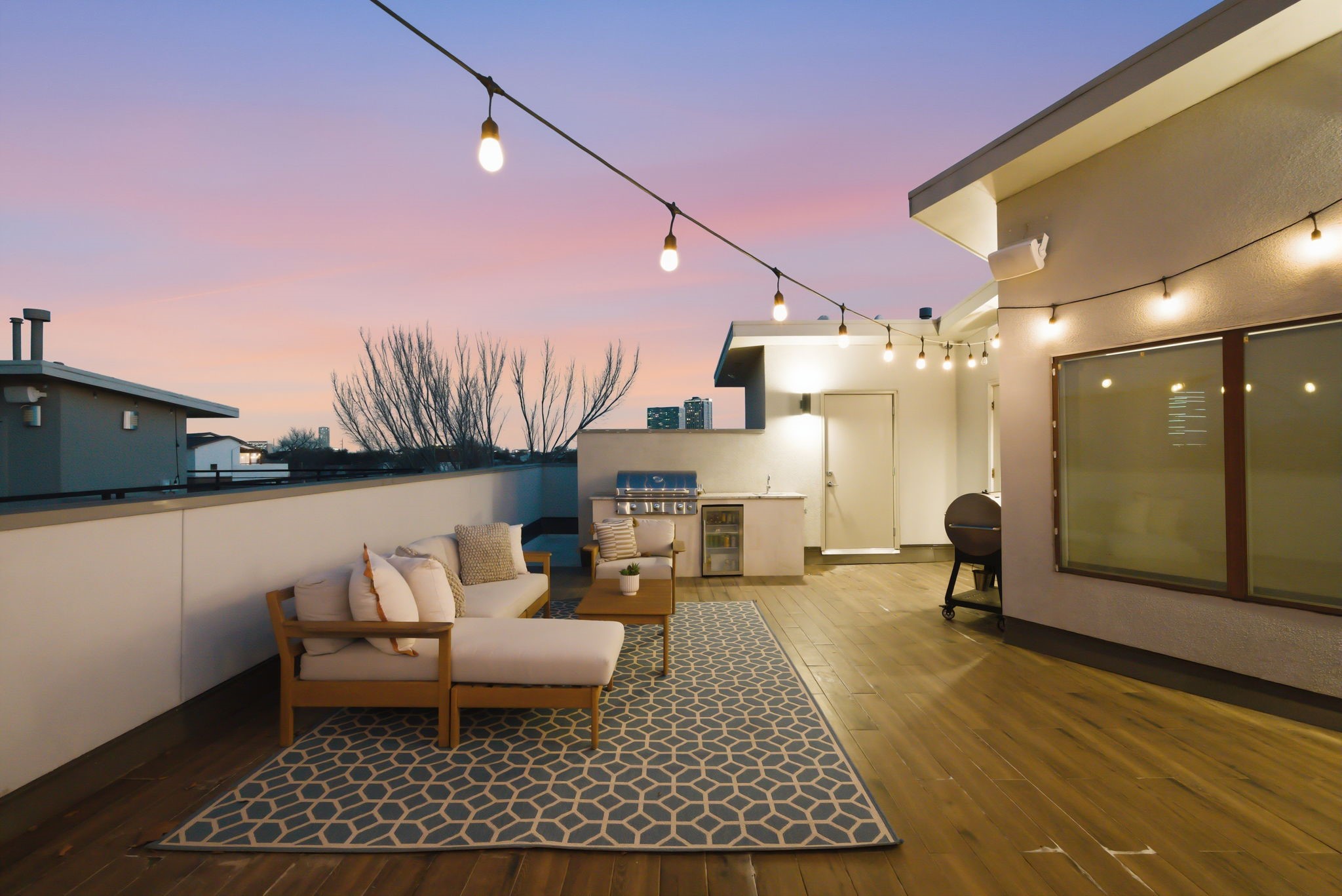 1405 Vermont Street, Unit A Houston, TX 77006 - Photo 37 of 46 Glow of string lights illuminates this expansive rooftop deck, creating a sophisticated setting for evening gatherings. Low-maintenance wood-look flooring leads to a high-end summer kitchen, while the spacious layout comfortably accommodates a plush lounge area for enjoying sunset views over the city. A designer geometric rug and sleek furniture enhance the luxury contemporary atmosphere of this private Bradford Mews retreat.