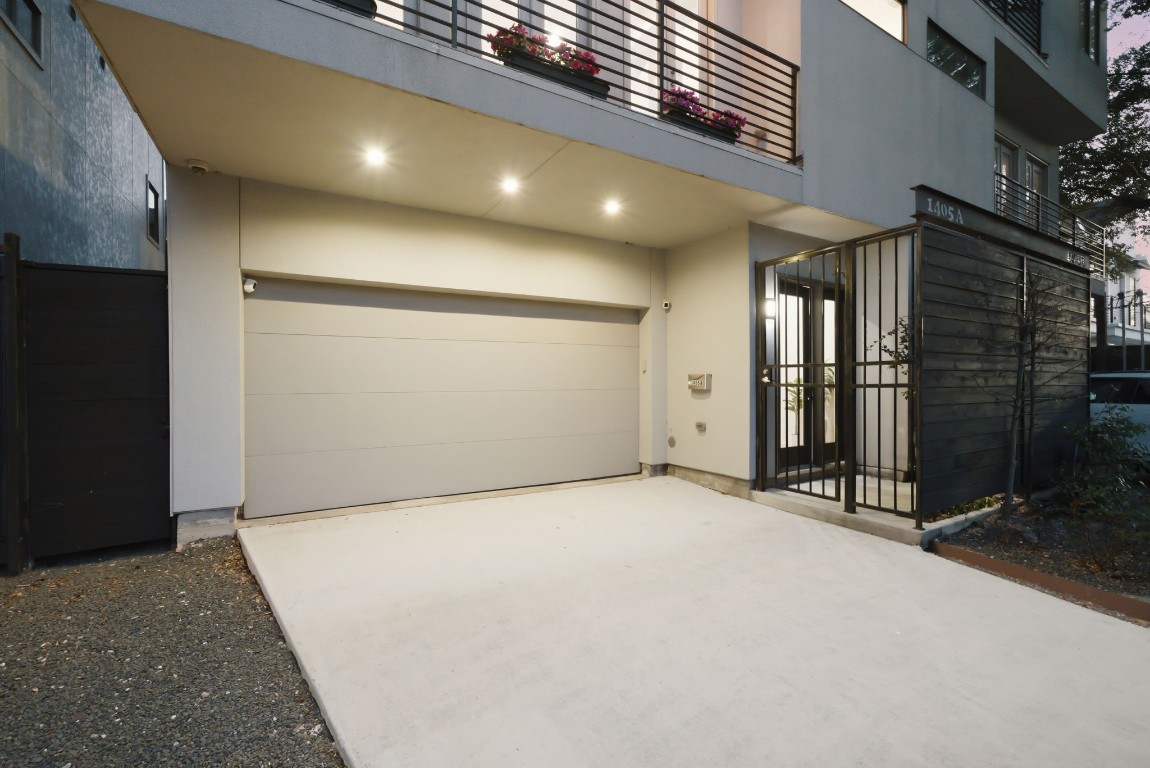 1405 Vermont Street, Unit A Houston, TX 77006 - Photo 44 of 46 Private driveway leads to a modern two-car garage at 1405 Vermont Street #A, featuring EV charging capability and sleek contemporary lines. Recessed soffit lighting illuminates the low-maintenance concrete approach, while a gated side entrance with black steel bars provides secure access to the front door. This sophisticated entryway perfectly complements the stucco and siding exterior of this luxury Inner Loop residence.
