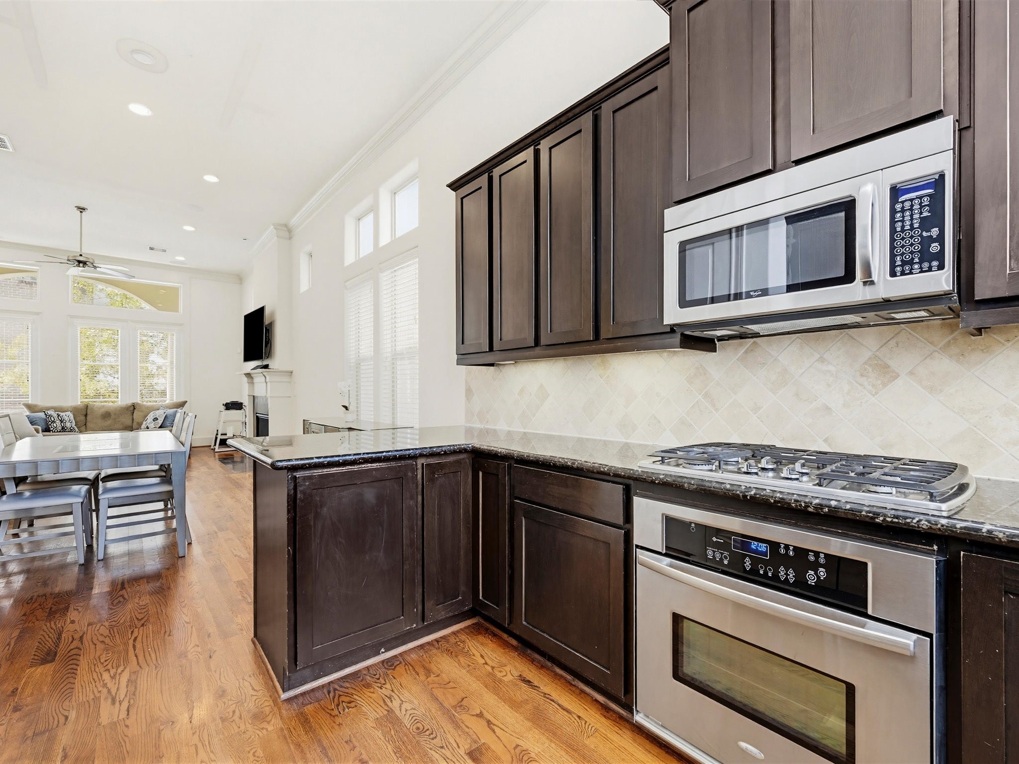 9427 London Bridge Station Houston, TX 77045 - Photo 11 of 30 This photo showcases a modern kitchen with dark wood cabinets, granite countertops, and stainless steel appliances, including a built-in microwave and gas cooktop. The kitchen opens to a bright dining and living area with large windows, hardwood floors, and a ceiling fan, creating a spacious and inviting atmosphere.
