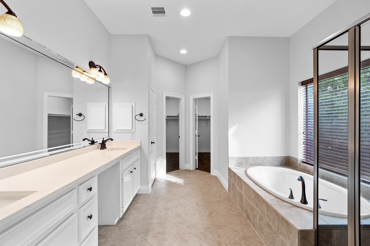 26411 Ridgefield Park Lane Cypress, TX 77433 - Photo 27 of 50 This primary bathroom features a spacious layout with a large vanity and dual sinks, a soaking tub with tile surround, and a separate glass-enclosed shower. Natural light streams through a large window with blinds, and the neutral tile floor complements the elegant fixtures.