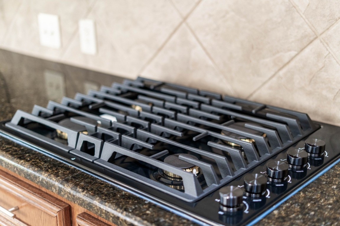 26411 Ridgefield Park Lane Cypress, TX 77433 - Photo 50 of 50 Modern kitchen with a sleek gas stovetop set on a dark granite countertop, complemented by light beige tile backsplash, offering a stylish and functional cooking space.