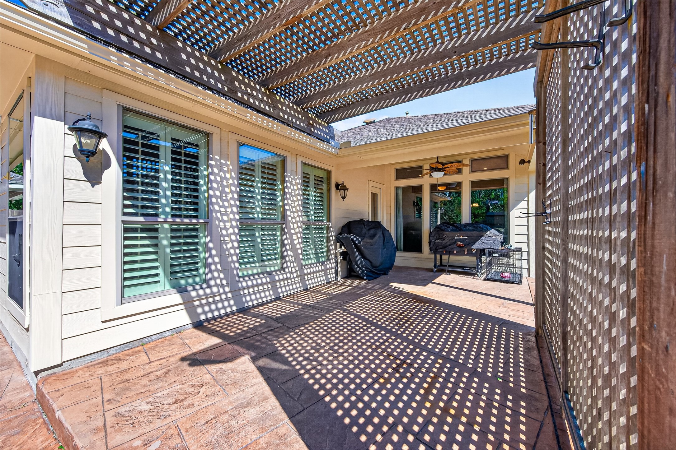 27443 Rebecca Field Lane Spring, TX 77386 - Photo 31 of 31 This photo shows a spacious outdoor patio with a pergola casting patterned shadows. It features large windows with shutters, outdoor lighting, and space for grilling and seating, offering a comfortable and stylish area for outdoor living.
