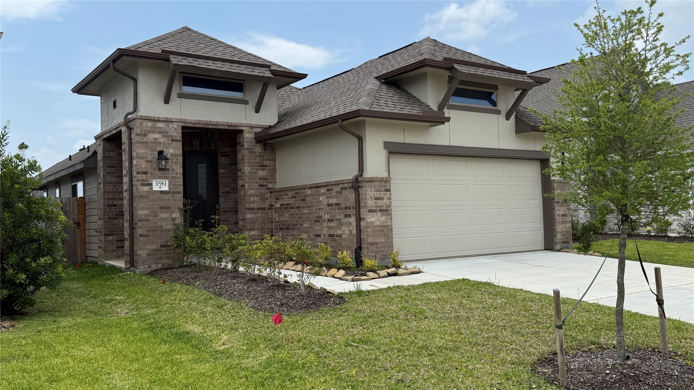 This photo showcases a modern single-story brick house with a two-car garage. It features a well-maintained front lawn, a small tree, and a neat garden bed lining the walkway to the front door.