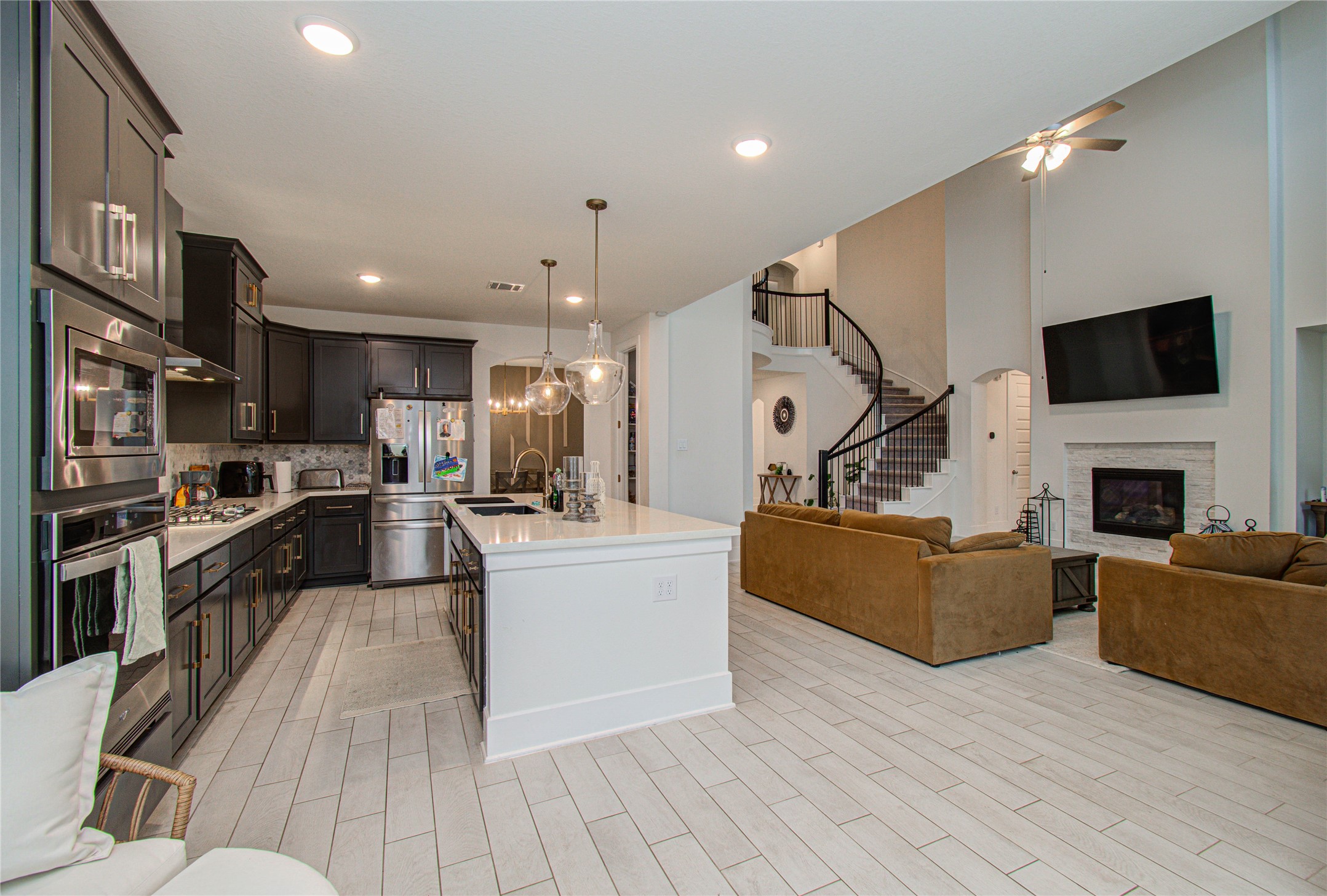 4211 Limestone Terrace Manvel, TX 77578 - Photo 9 of 33 This photo showcases a spacious, open-concept living area with a modern kitchen featuring dark cabinetry and stainless steel appliances. The adjoining living room offers a cozy fireplace and ample seating. A stylish staircase leads to an upper level, enhancing the home's elegant design.